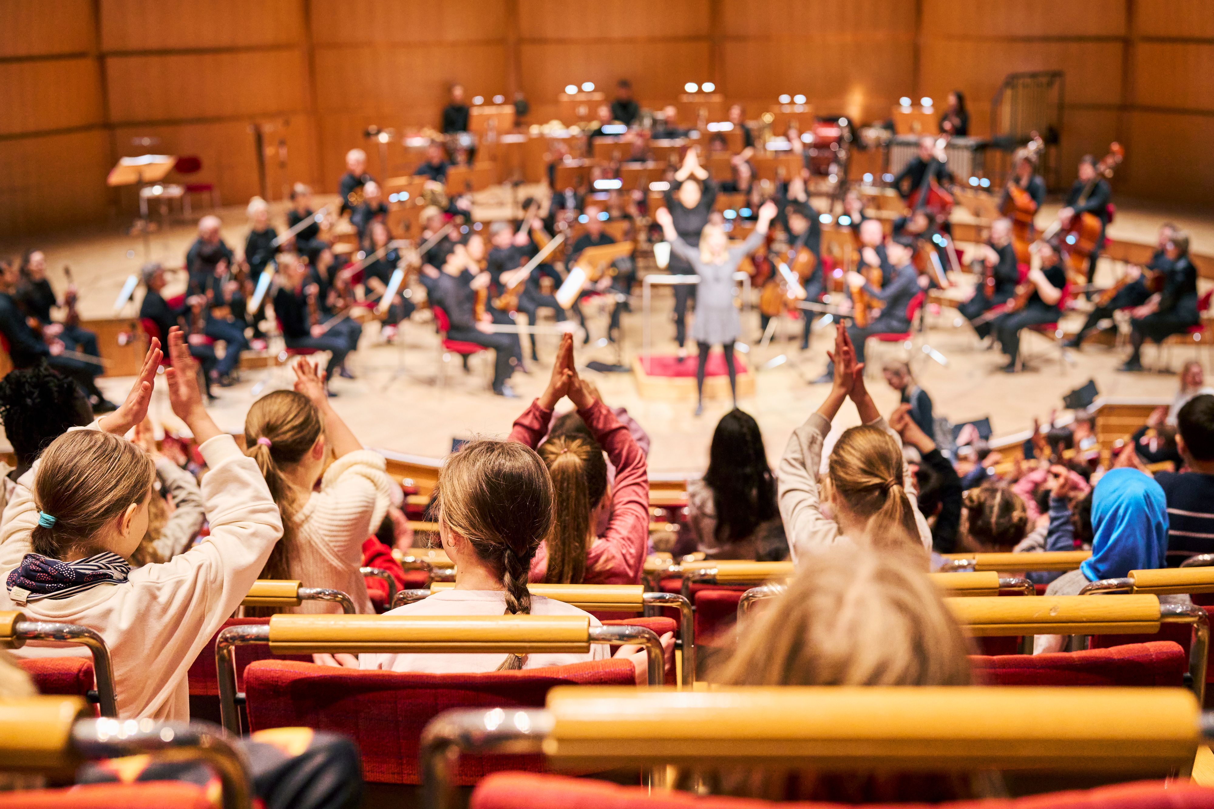 Kinder sitzen in den Besucherreihen der Kölner Philhramonie und klatschen in die Hände, auf der Bühne steht ein junges Mädchen, hinter ihm Musikerinnen und Musiker des Gürzenich-Orchesters