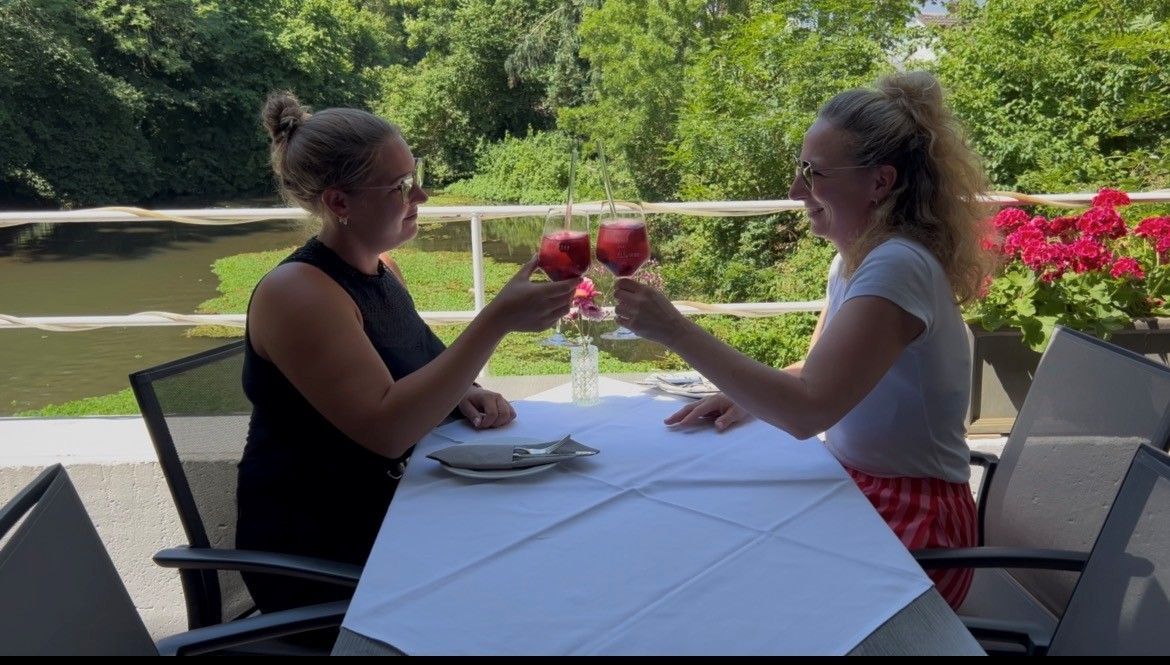 Auf dem Bild sind zwei Frauen an einem Tisch auf einer Terrasse zu sehen, die sich zuprosten.