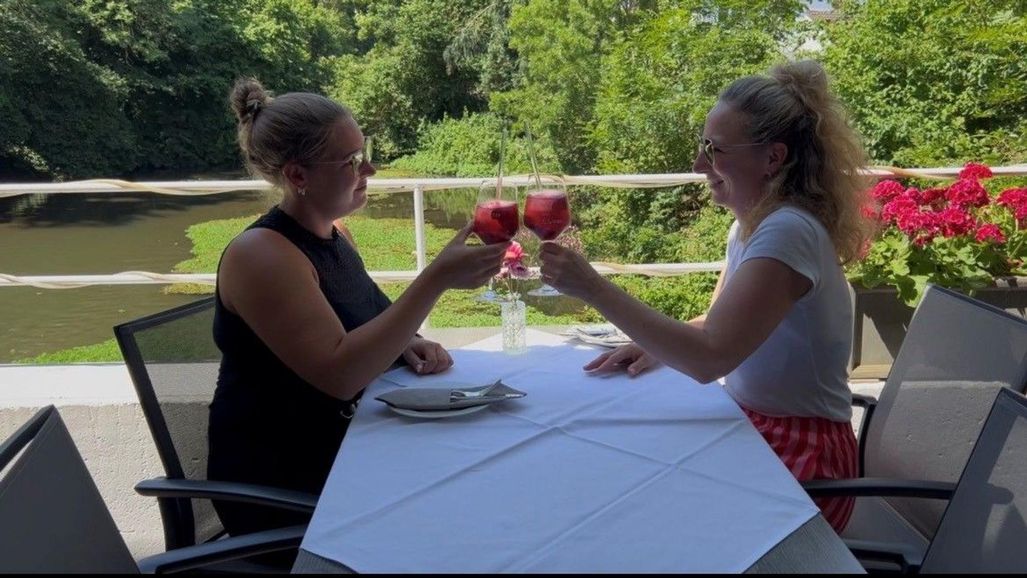 Auf dem Bild sind zwei Frauen an einem Tisch auf einer Terrasse zu sehen, die sich zuprosten.