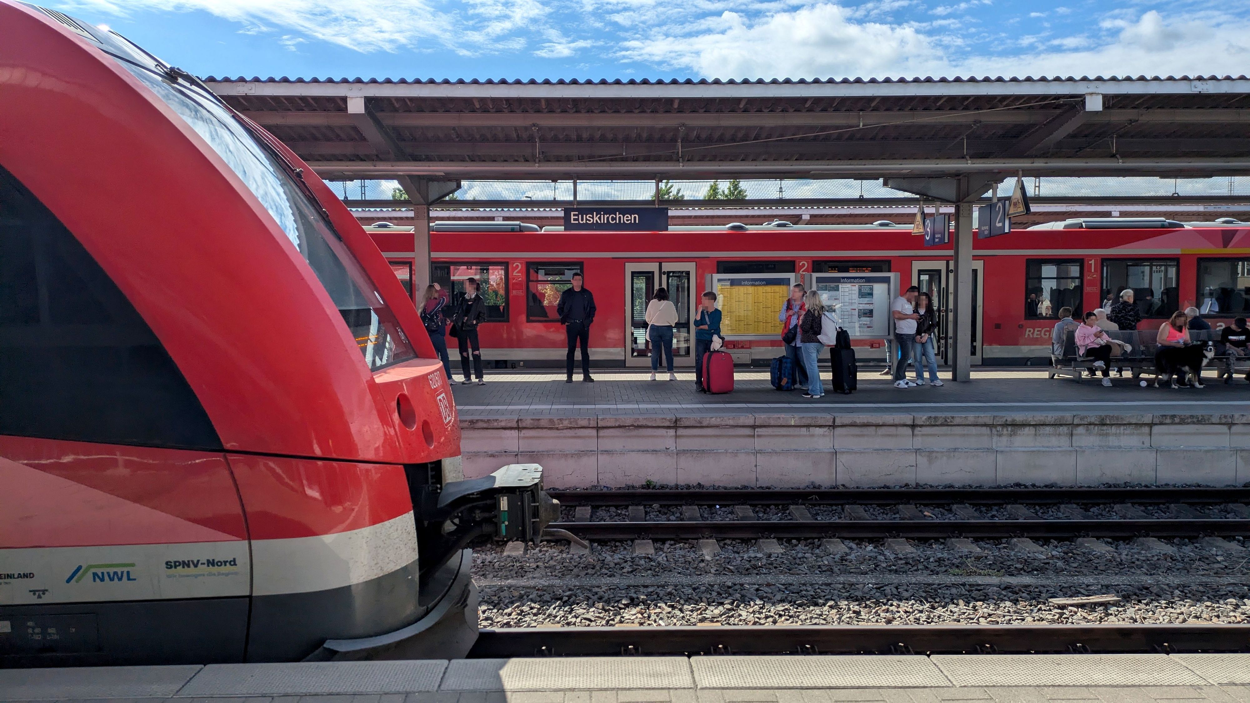 Ein roter Vareo-Triebwagen fährt auf Gleis 1 im Bahnhof Euskirchen ein. Dahinter sind wartende Reisende und ein weiterer Regionalzug zu sehen.