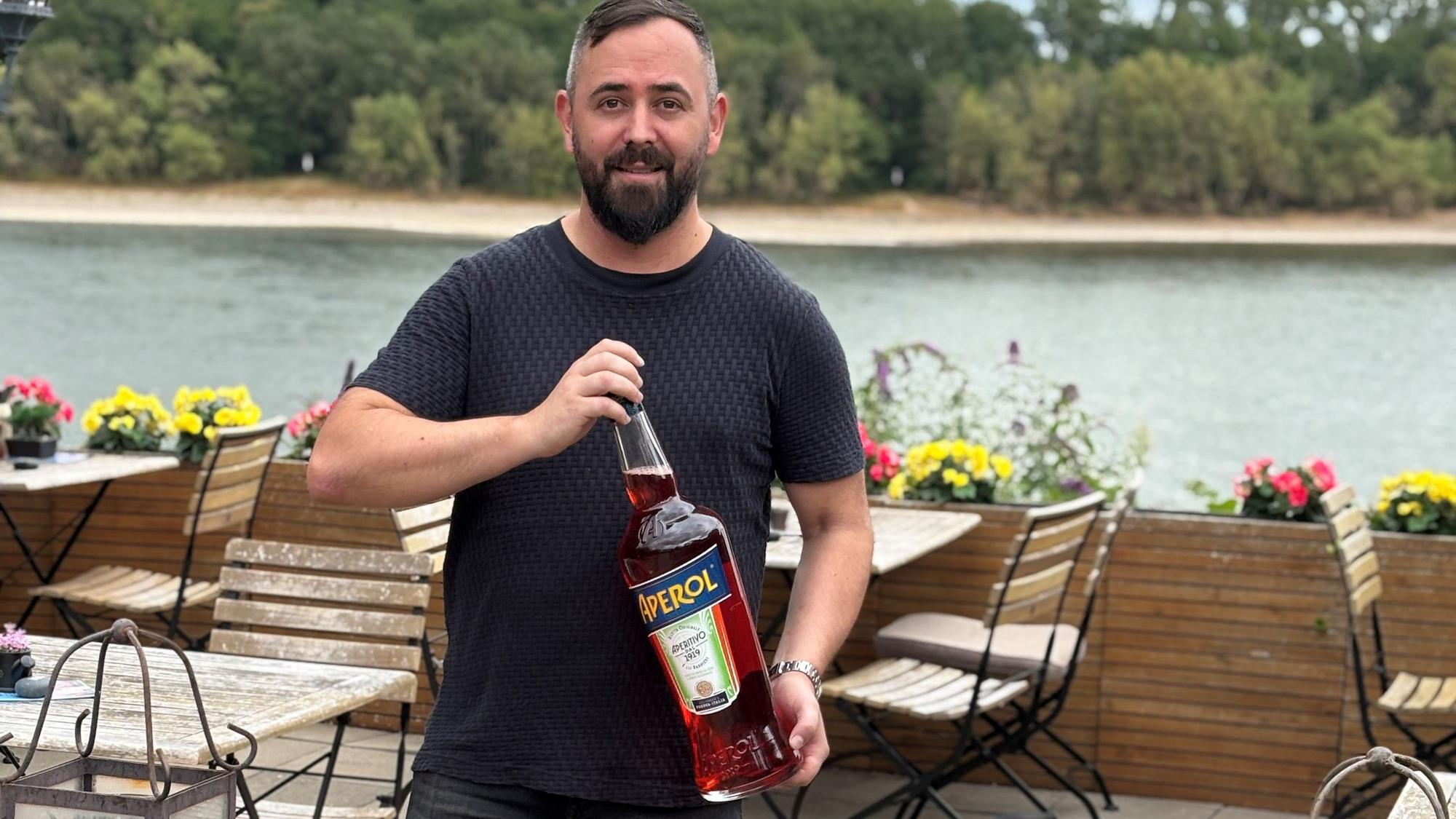 Auf dem Bild ist ein Mann zu sehen, der eine große Flasche Aperol in den Händen hält.