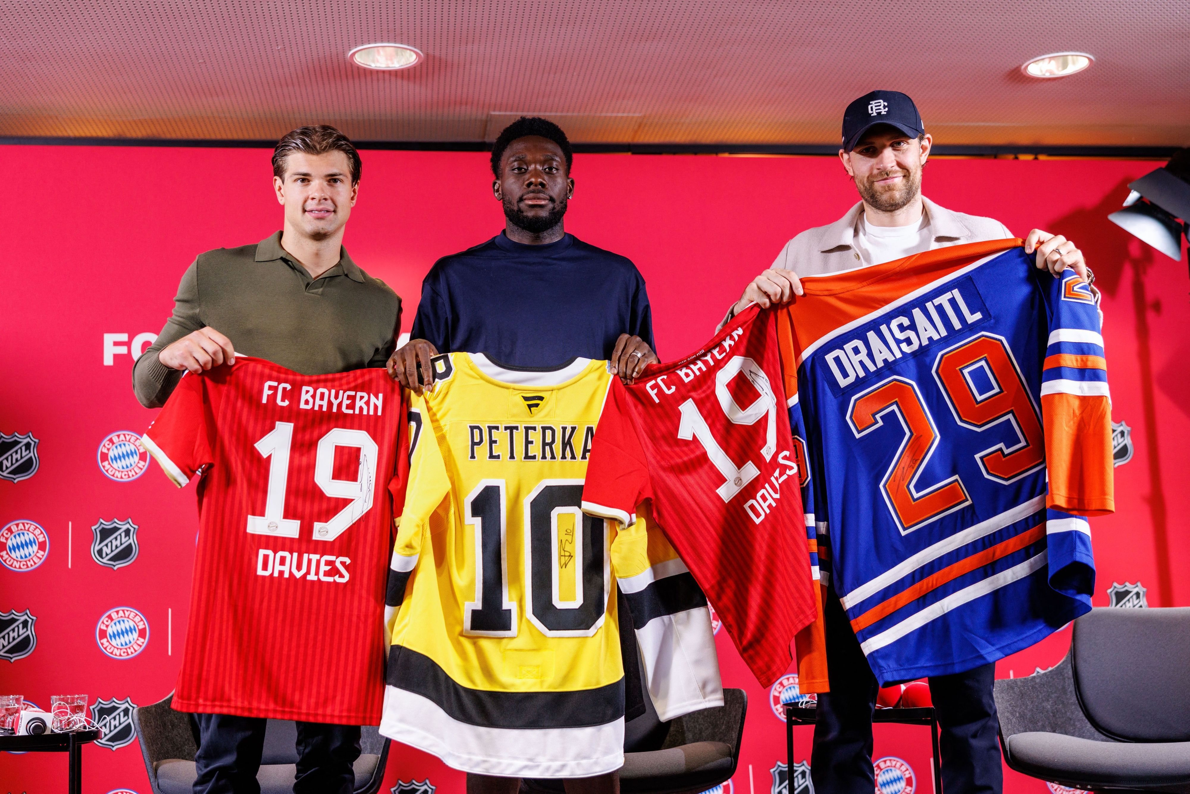 20.08.2026, Bayern, München: Eishockey, Fußball: Gemeinsame Pk des FC Bayern München und der National Hockey League (NHL). John-Jason Peterka (l-r), Spieler der Boston Bruins, Alphonso Davies, Spieler vom FC Bayern München, und Leon Draisaitl, Spieler der Edmonton Oilers, posieren auf der Pressekonferenz mit Trikots. Foto: Matthias Balk/dpa - WICHTIGER HINWEIS: Gemäß den Vorgaben der Bundesliga bzw. des DFB Deutscher Fußball-Bund ist es untersagt, in dem Stadion und/oder vom Spiel angefertigte Fotoaufnahmen in Form von Sequenzbildern und/oder videoähnlichen Fotostrecken zu verwerten bzw. verwerten zu lassen. +++ dpa-Bildfunk +++