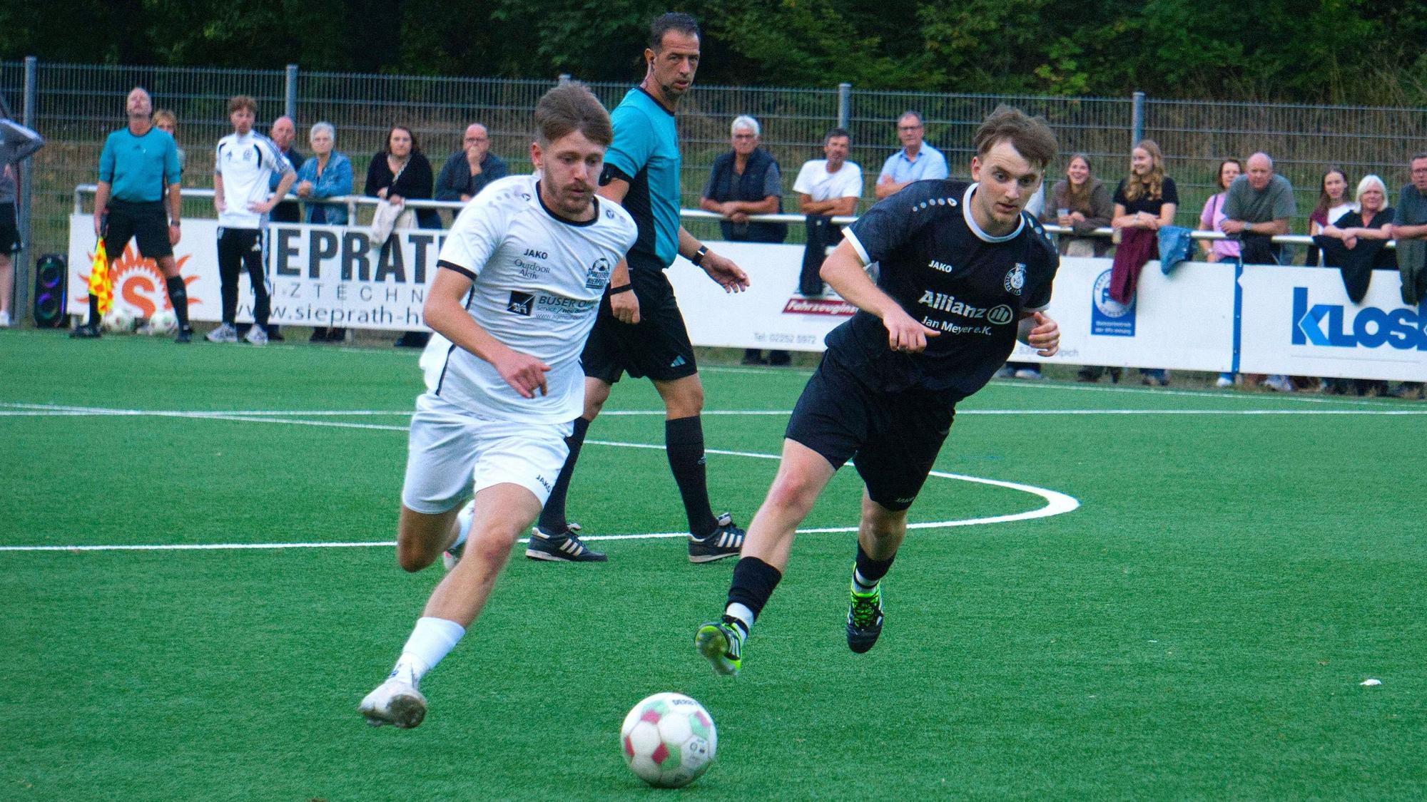 Zwei Fußballer im Sprintduell um den Ball, der Schriedsrichter schaut im Hintergrund zu.
