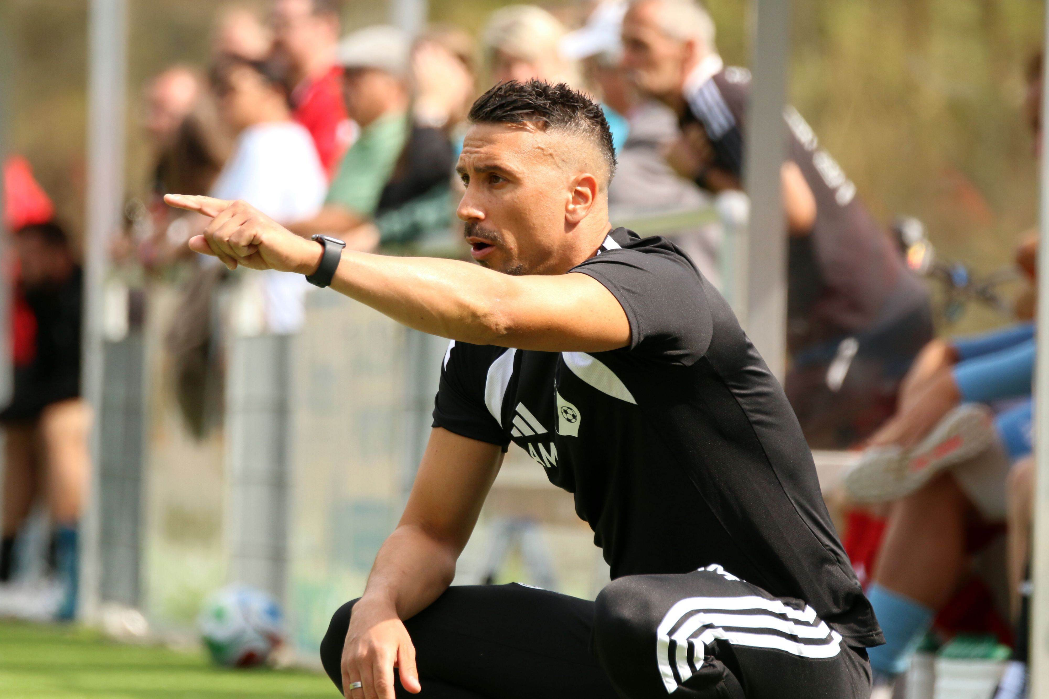 Angelo Mazza hockt neben dem Spielfeld und zeigt mit ausgestrecktem Zeigefinger etwas an.