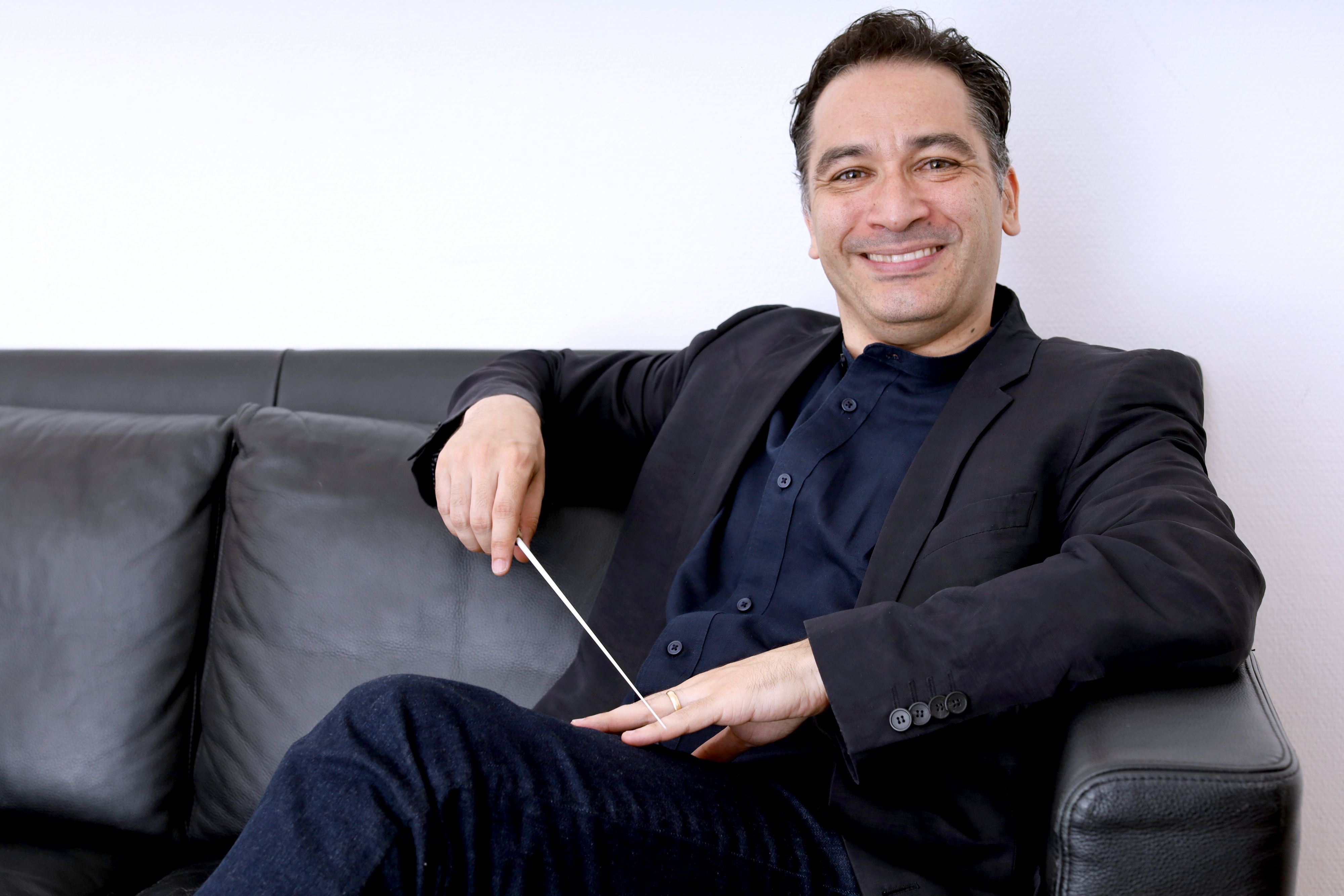 Andrés Orozco-Estrada (Jahrgang 1977), seit der Spielzeit 2025/26 Generalmusikdirektor der Stadt Köln und Kapellmeister des Gürzenich-Orchesters, sitzt mit mit seinem Dirigierstock auf einem schwarzen Ledersofa und lächelt herzlich.