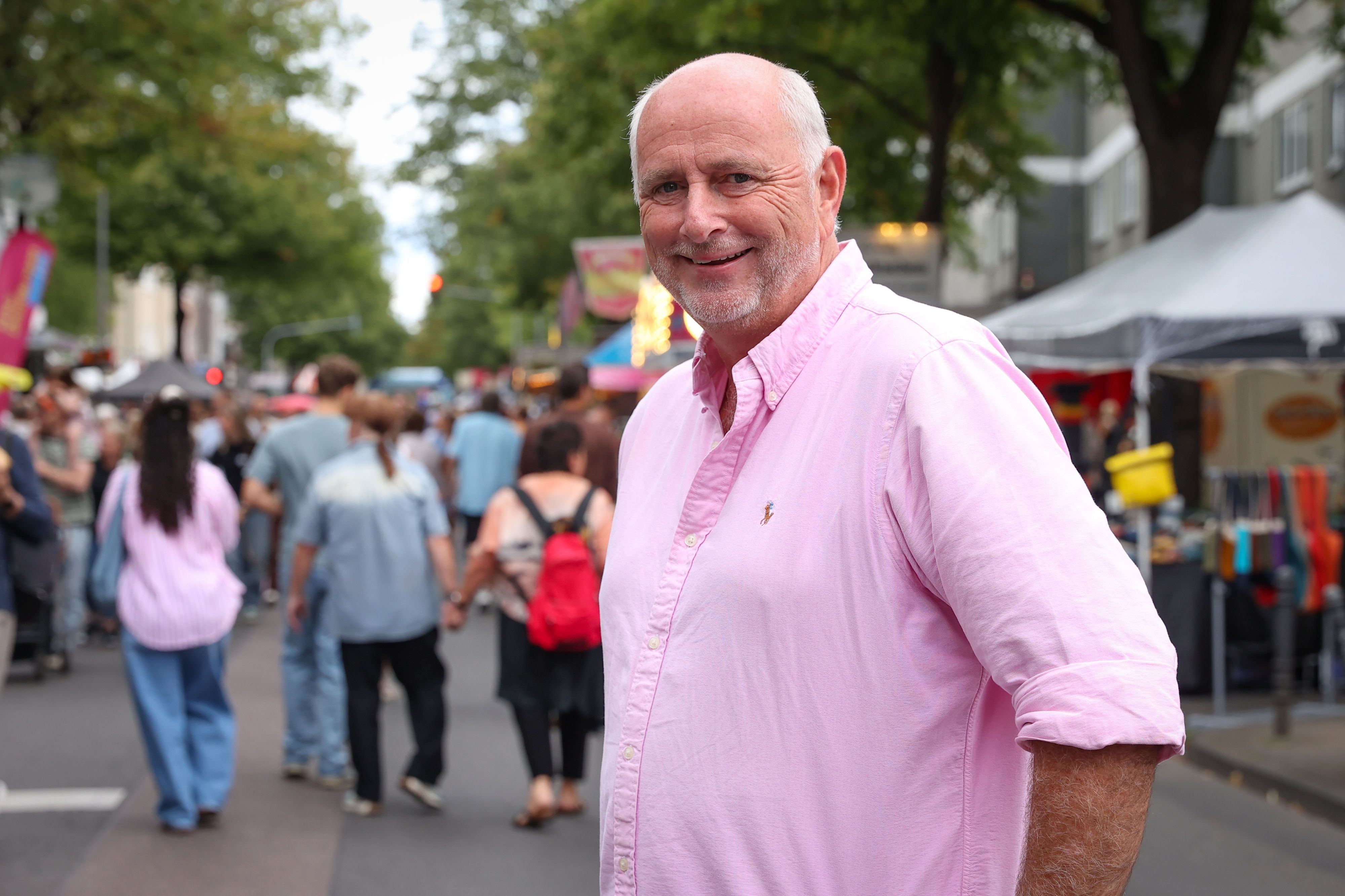 Ein Mann mit Bart und rosa Shirt steht auf einer Straße, auf der Stände aufgebaut sind und viele Menschen unterwegs sind.