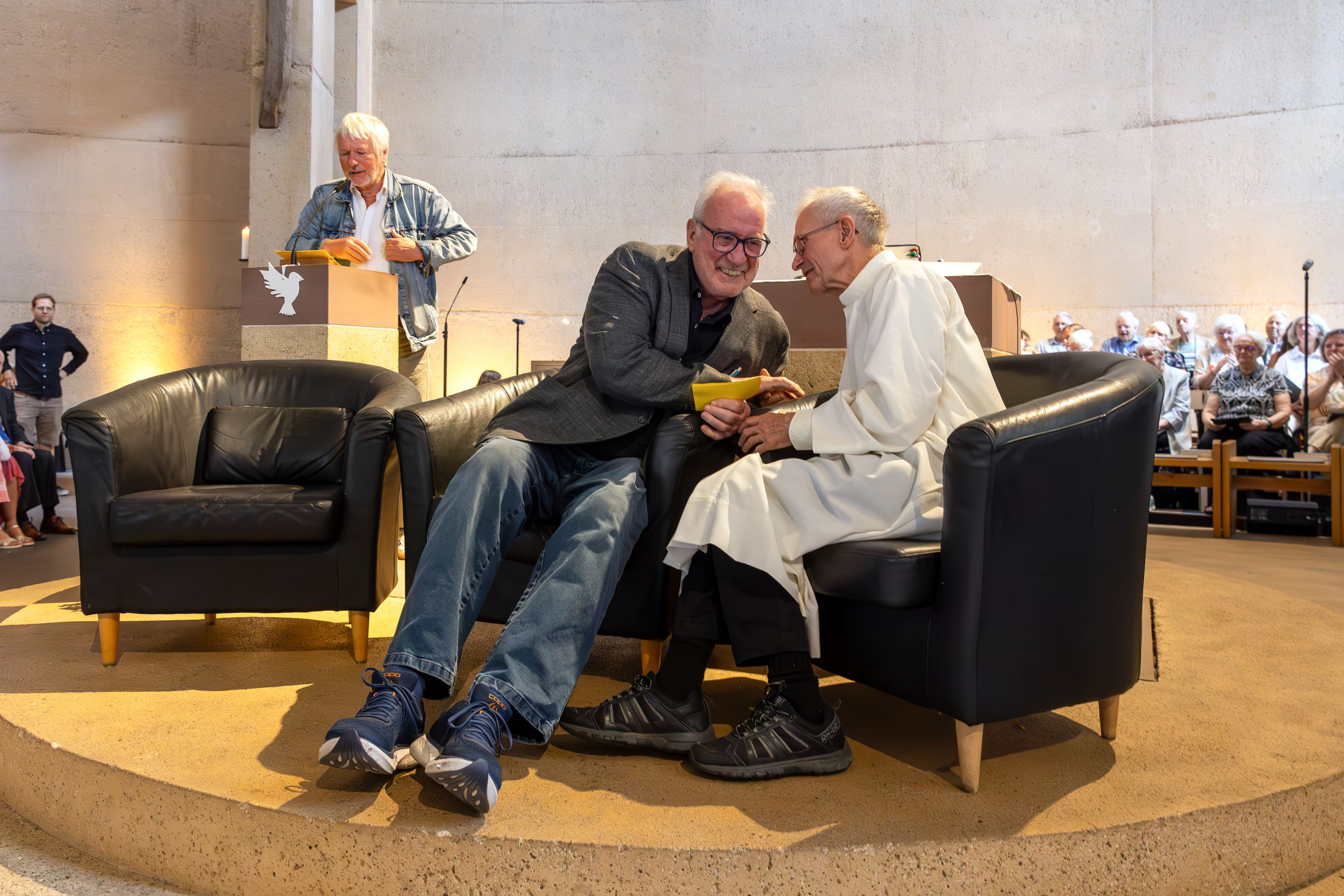 Ein Mann steht am Ambo im Altarraum einer Kirche, auf Sesseln im Vordergrund sitzen zwei Herren, einer trägt die Albe, das Messgewand des Priesters.