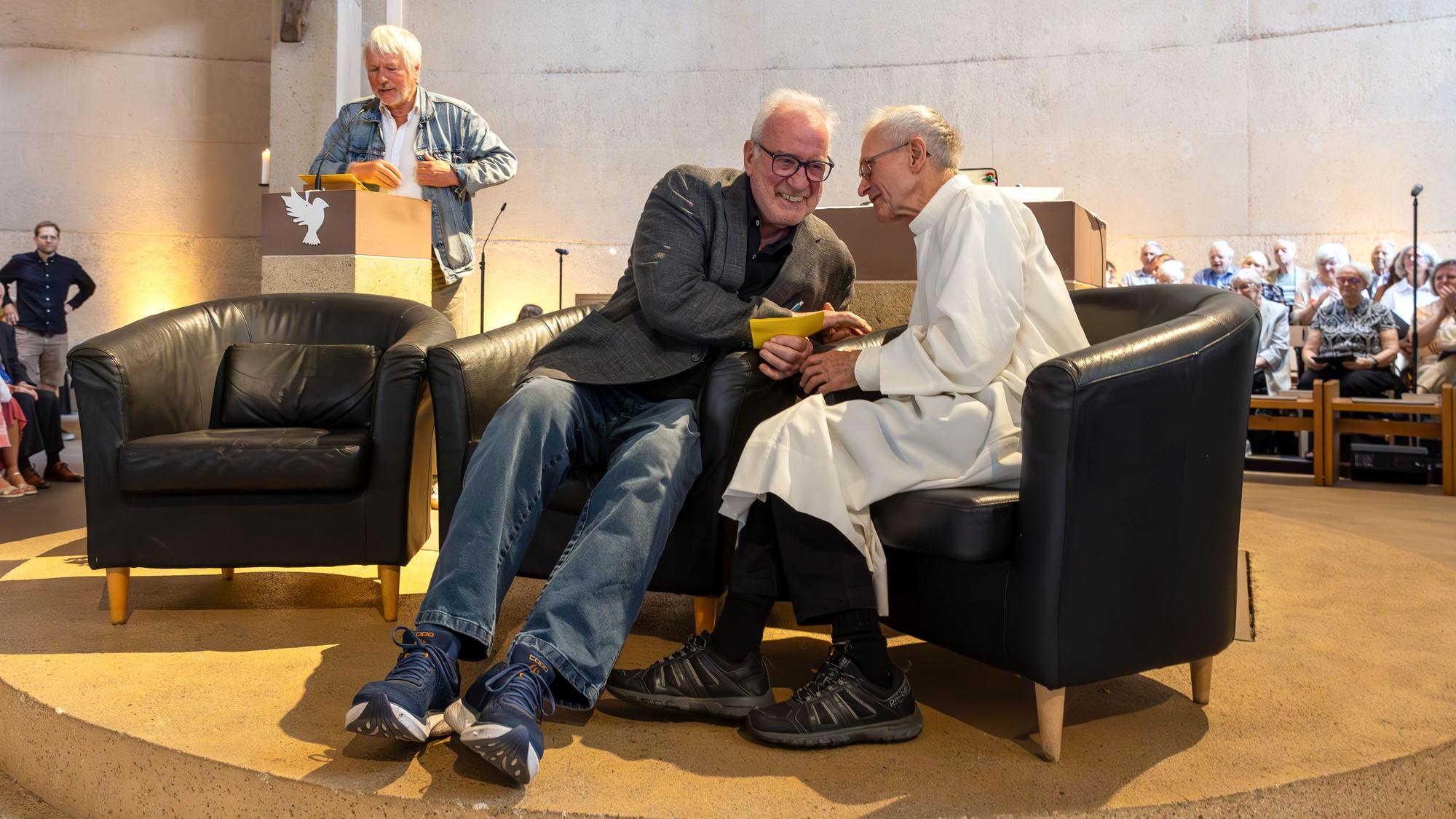 Ein Mann steht am Ambo im Altarraum einer Kirche, auf Sesseln im Vordergrund sitzen zwei Herren, einer trägt die Albe, das Messgewand des Priesters.