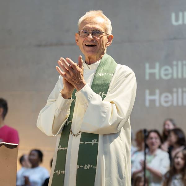 Der Kölner Pfarrer Franz Meurer im Priestergewand klatscht in die Hände. Hinter ihm ist ein Chor zu sehen und Buchstaben an der Wand, die den Text des Liedes zeigen.