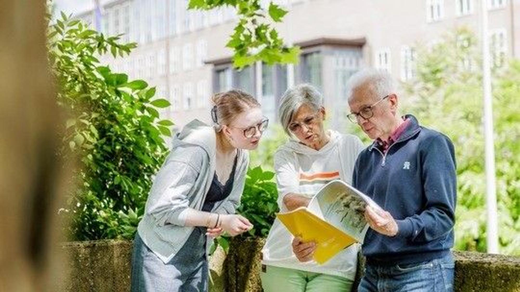 ZWei Seniorenstudenten und eie junge Frau schauen in eine Broschüre, im Hintergrund sieht man das Hauptgebäude der Uni Köln.