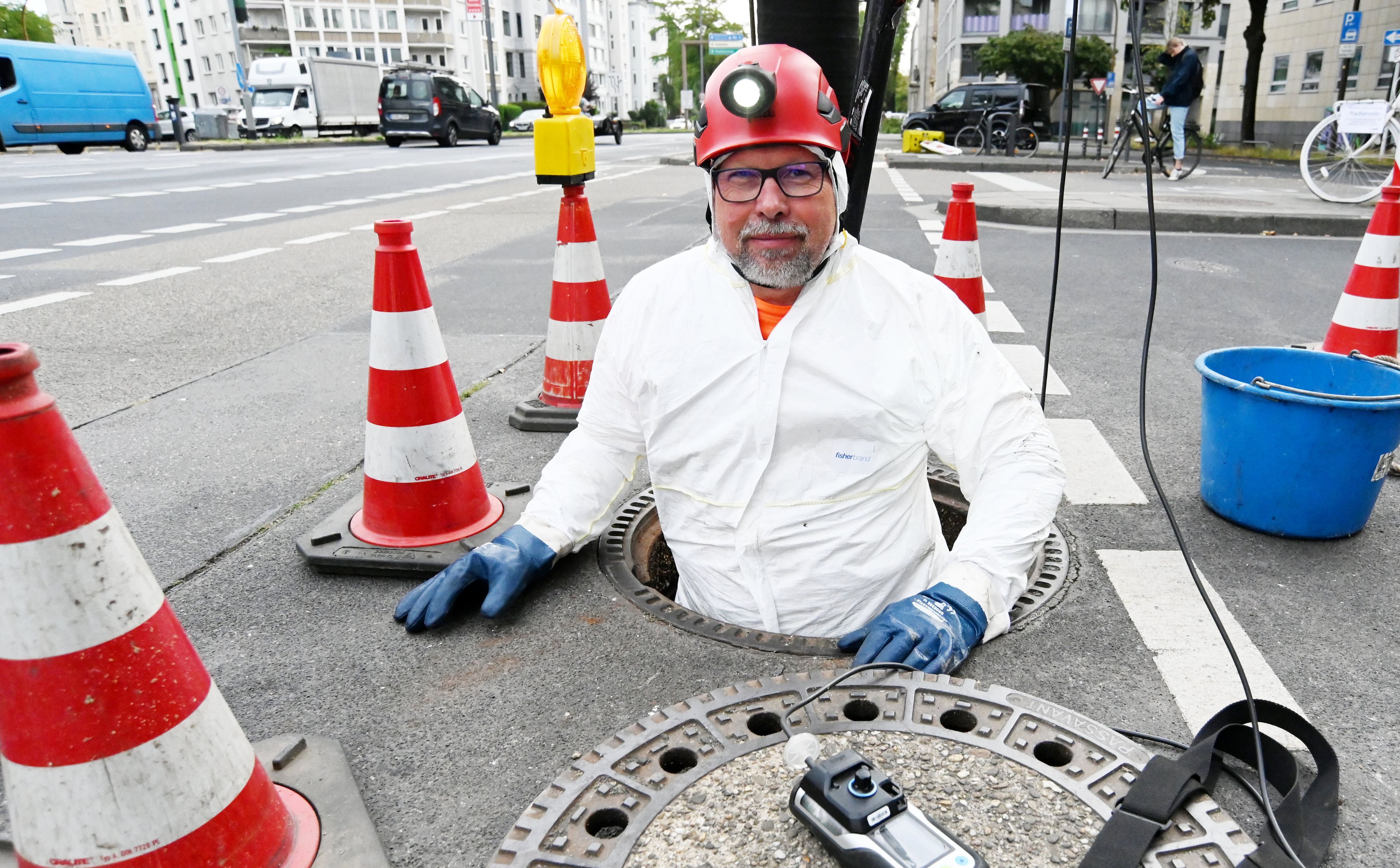 Ein Mann mit Helm und im weißen Schutzanzug befindet sich bis zum Bauch in einem Schacht zur Kanalisation.
