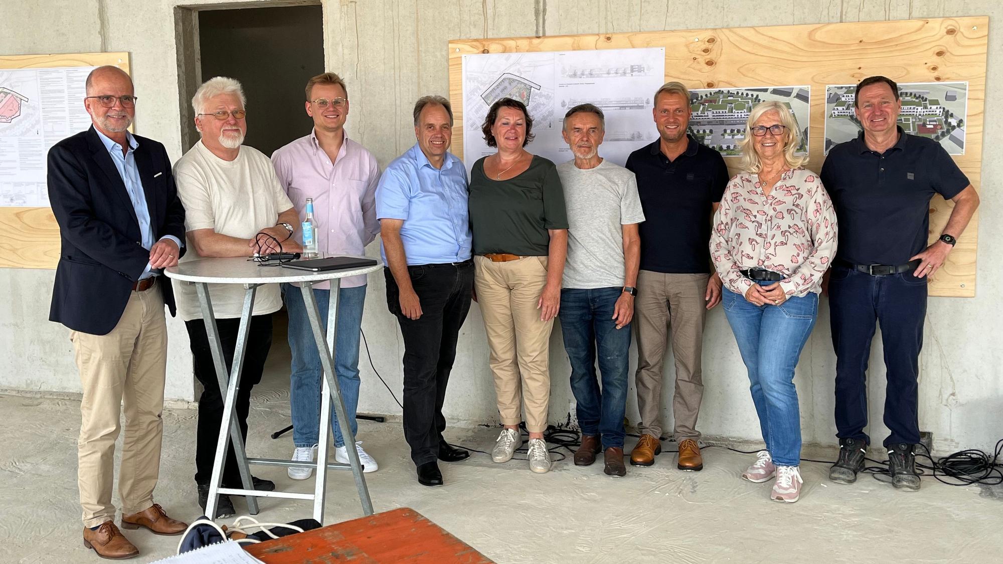 Männer und Frauen stehen nebeneinander an einer Wand, an der Baupläne befestigt sind. Zwei Männer stehen an einem Stehtisch. Sie befinden sich in einem Rohbau.