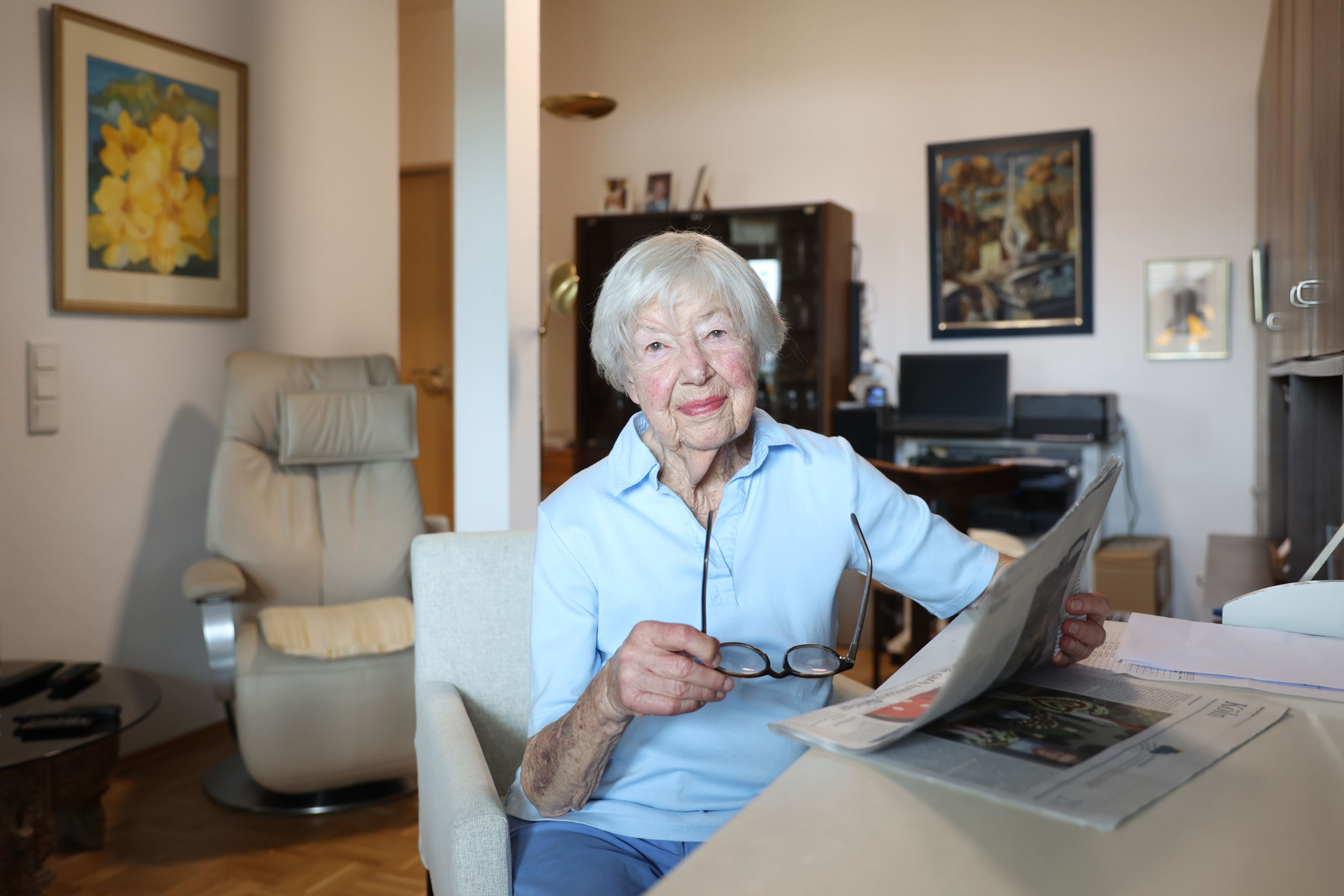 Helga Schmitt sitzt in ihrem Wohnzimmer am Tisch, in der Hand eine Lesebrille und eine Zeitung. Sie schaut freundlich in die Kamera.