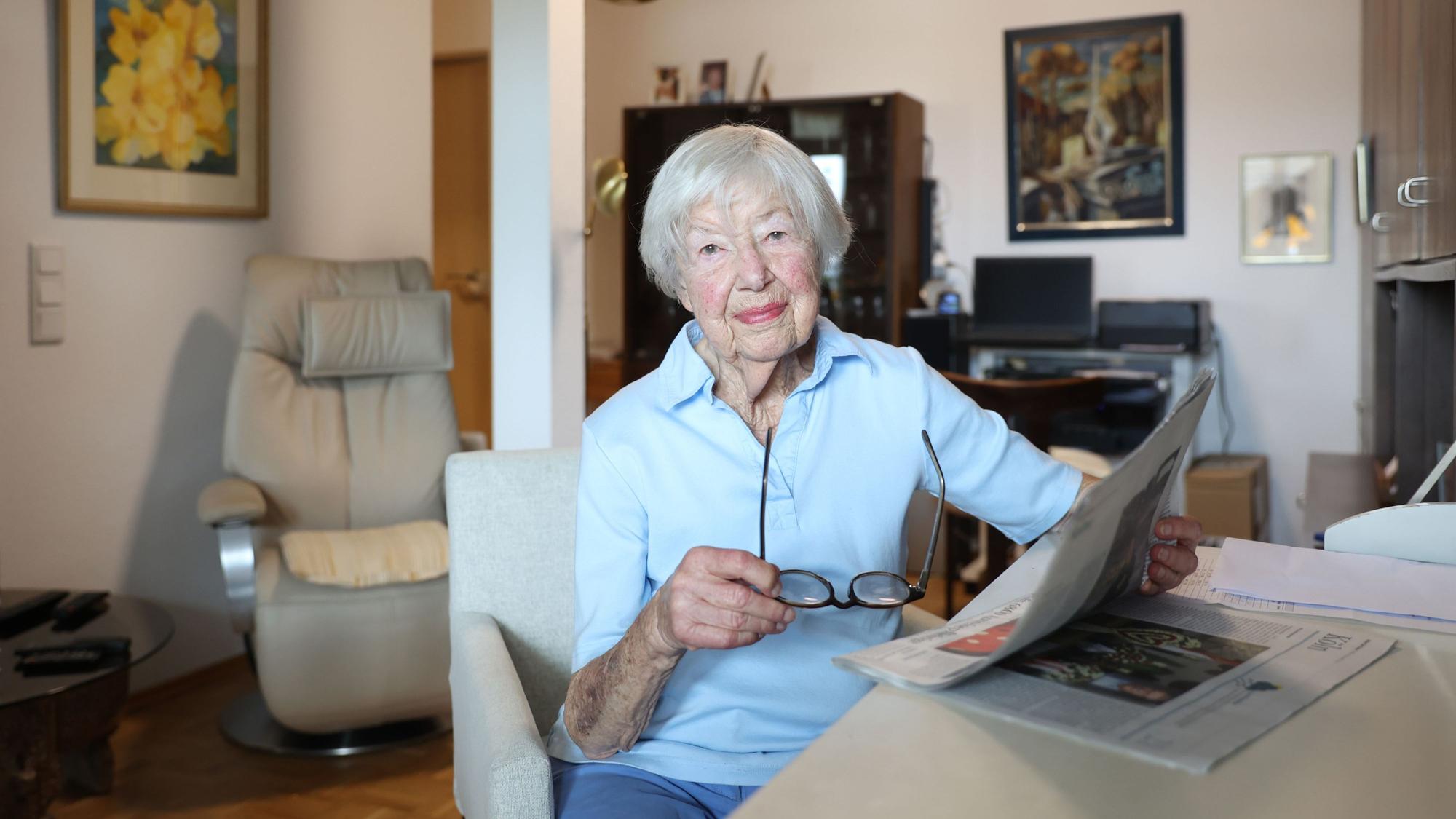 Helga Schmitt sitzt in ihrem Wohnzimmer am Tisch, in der Hand eine Lesebrille und eine Zeitung. Sie schaut freundlich in die Kamera.