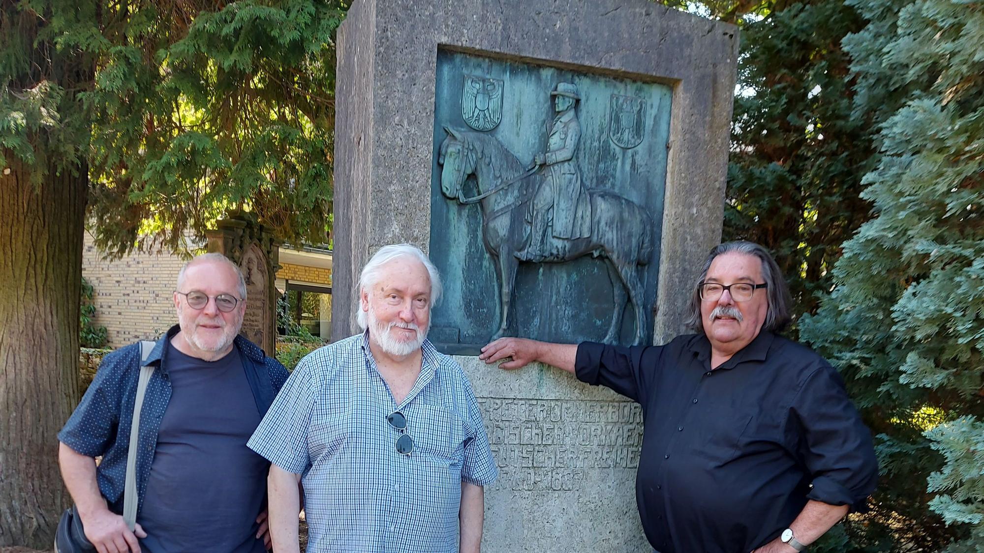 Drei Männer stehen vor einem Denkmal mit Bronzeplakette. Darauf ein Mann zu Pferd.