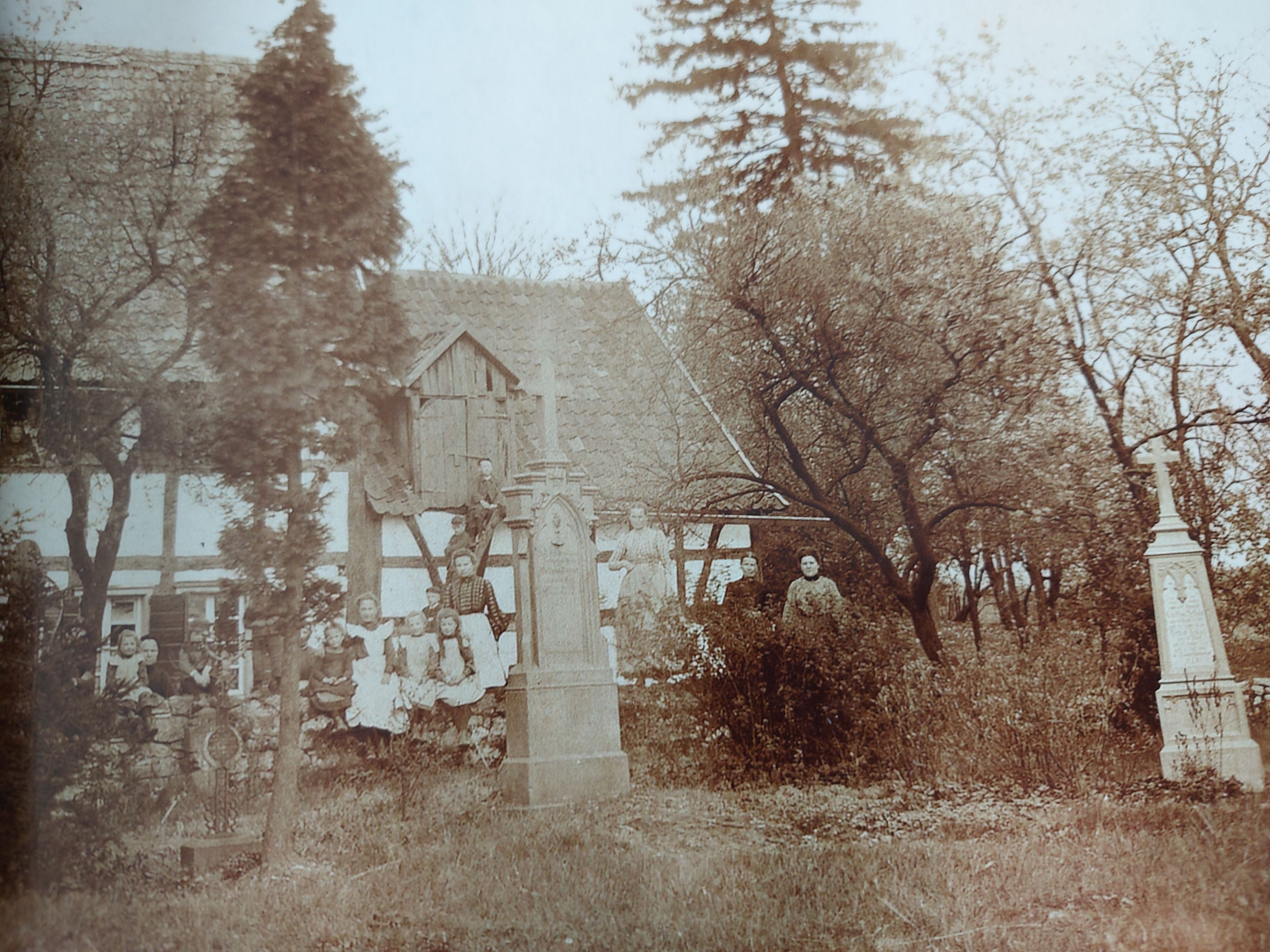 Ein historisches Foto um 1900 zeigt viele Menschen auf einem Friedhof mit Grabkreuzen. Dahinter ein Fachwerkhaus.