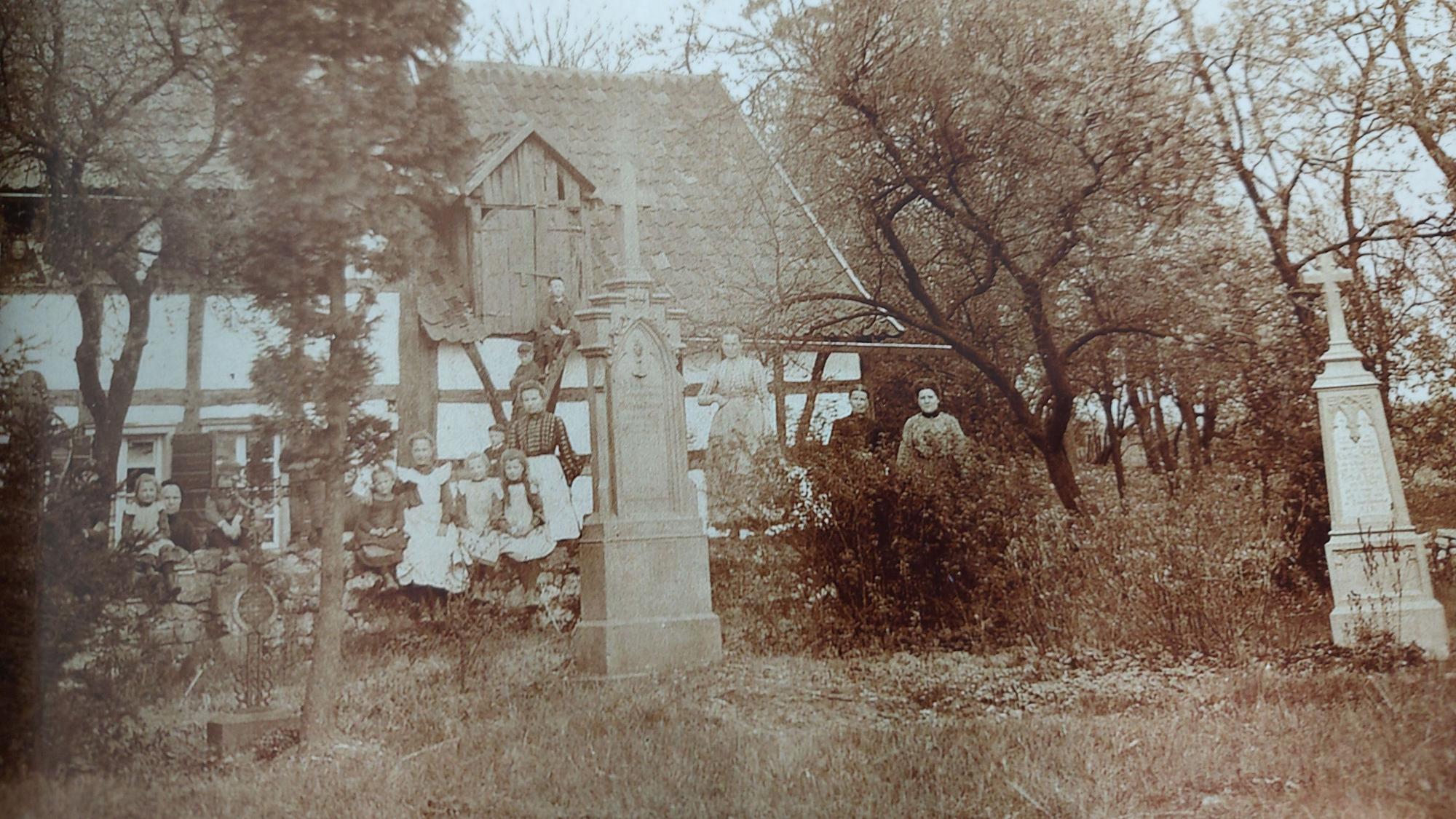 Ein historisches Foto um 1900 zeigt viele Menschen auf einem Friedhof mit Grabkreuzen. Dahinter ein Fachwerkhaus.