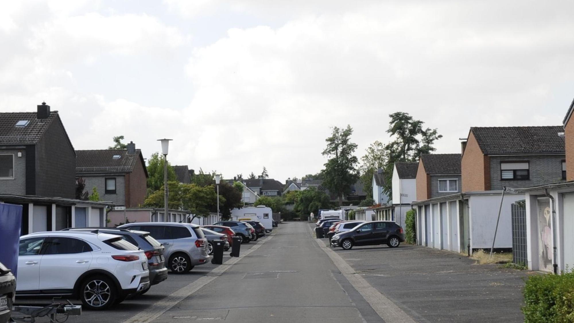 Zu sehen ist eine Straße mit Garagen und parkenden Autos davor, dahinter sind Häuser zu sehen.