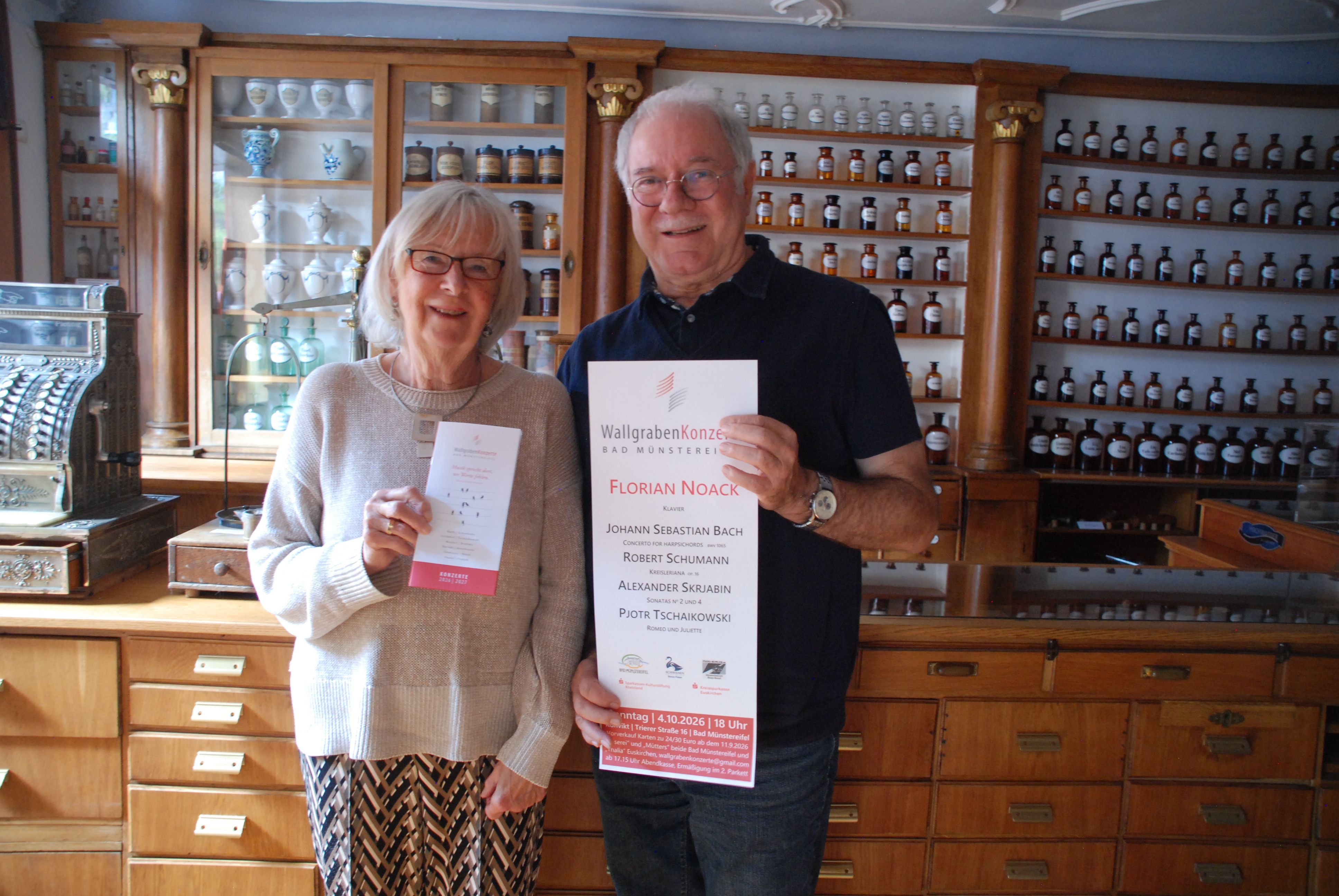 Eine Frau und ein Mann stehen vor einer Wand mit Apothekenfläschen und halten ein Plakat und einen Flyer in der Hand.
