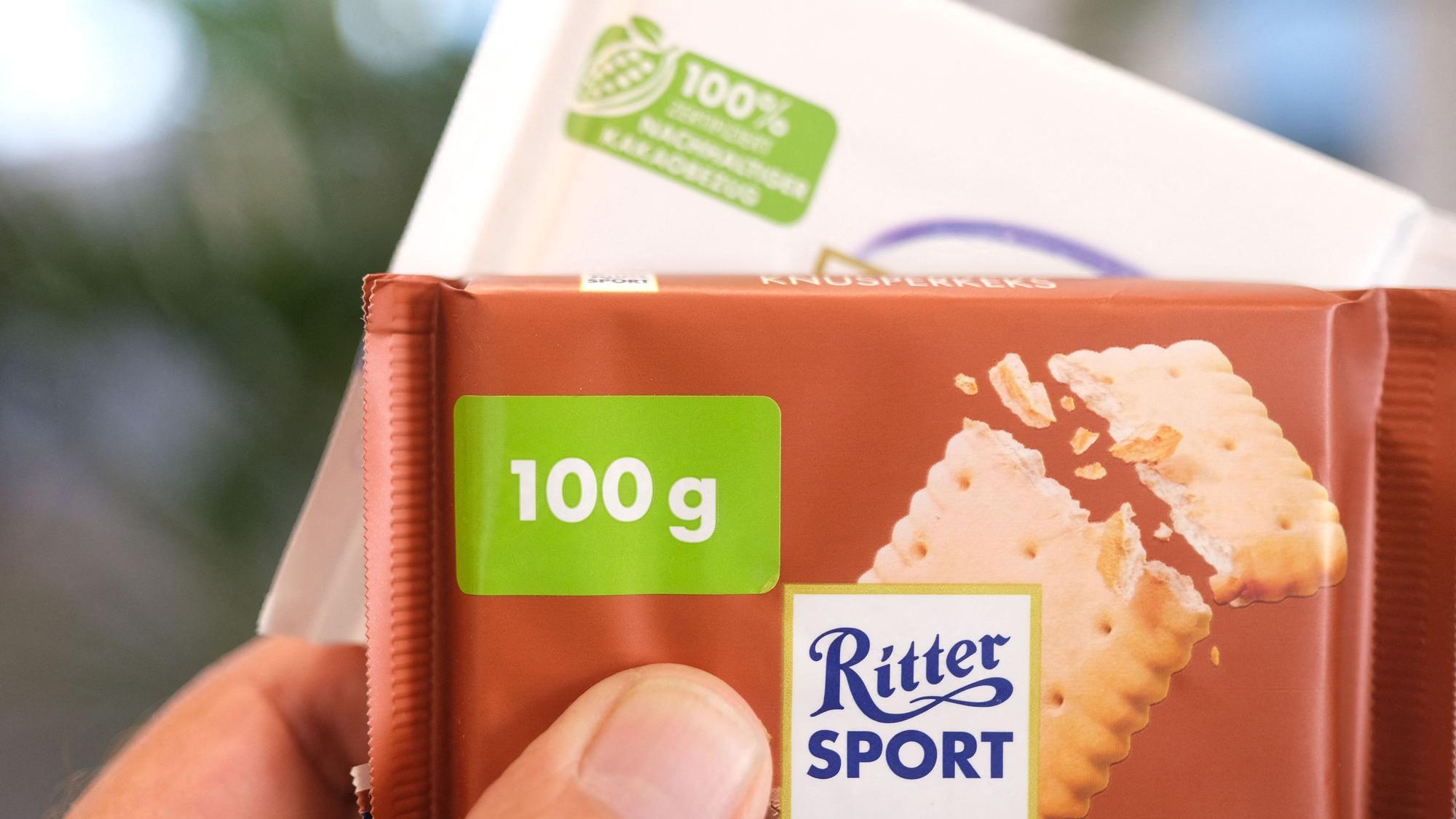 Eine Hand hält zwei Tafeln Schokolade von Ritter Sport. Auf der vorderen Tafel ist auf der Verpackung ein Hinweis mit einem Sticker überklebt.
