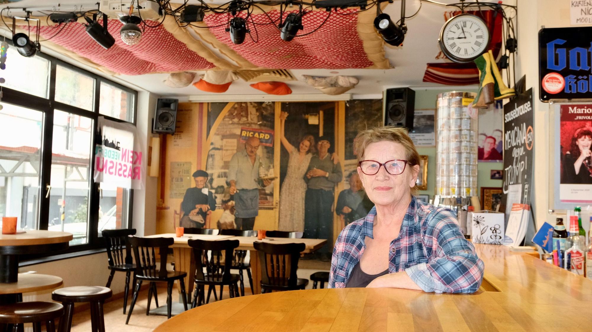 Eine Frau mit kurzem Haar, Brille und Karo-Hemd sitzt an der Bar in einer geräumigen Kneipe.