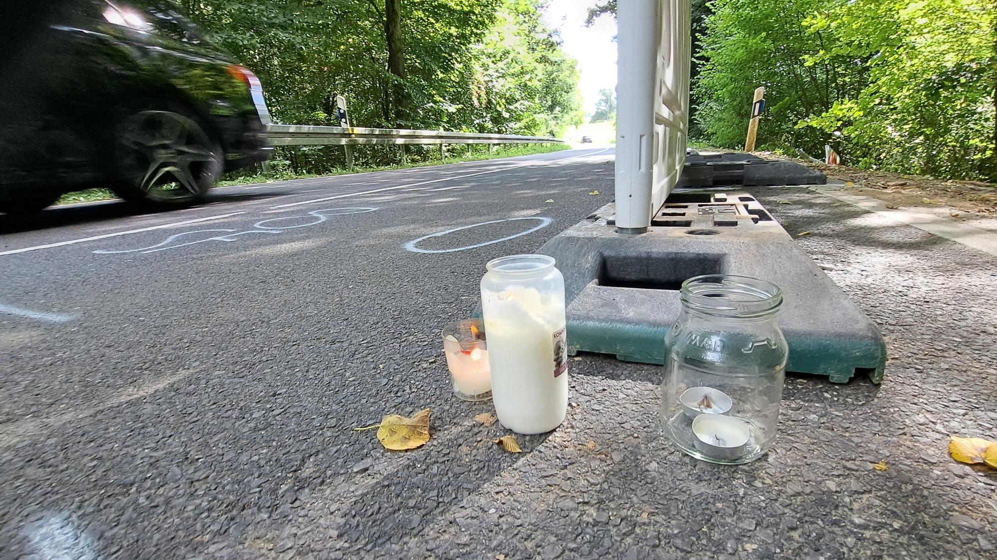 Eine Straße mit Grabkerzen rechts vor einer Bake und links einem vorbeifahrenden Auto.