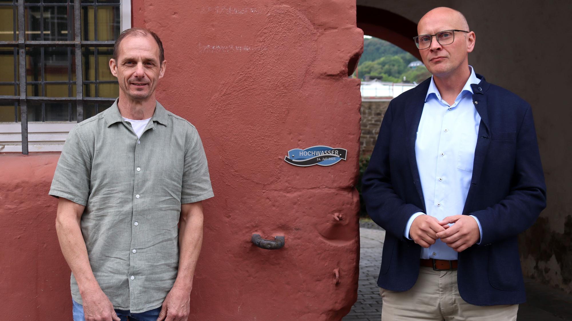 Bad Münstereifels Bürgermeister Sebastian Glatzel und Ralf Wassong stehen an der Flutmarke, die am Rathaus angebracht ist.