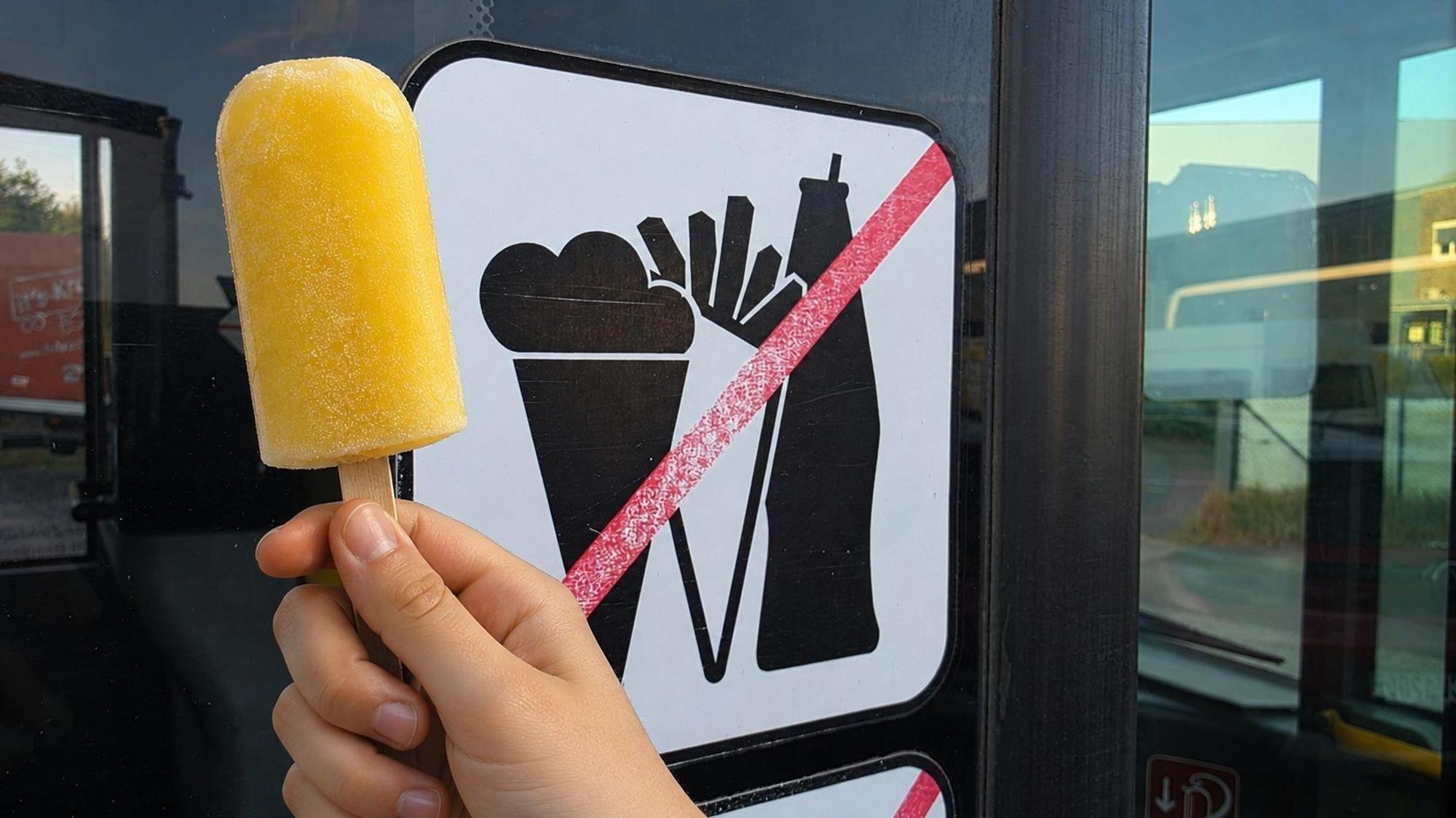 Eine Kinderhand hält ein Eis vor dem „Essen verboten“-Schild eines Linienbusses.