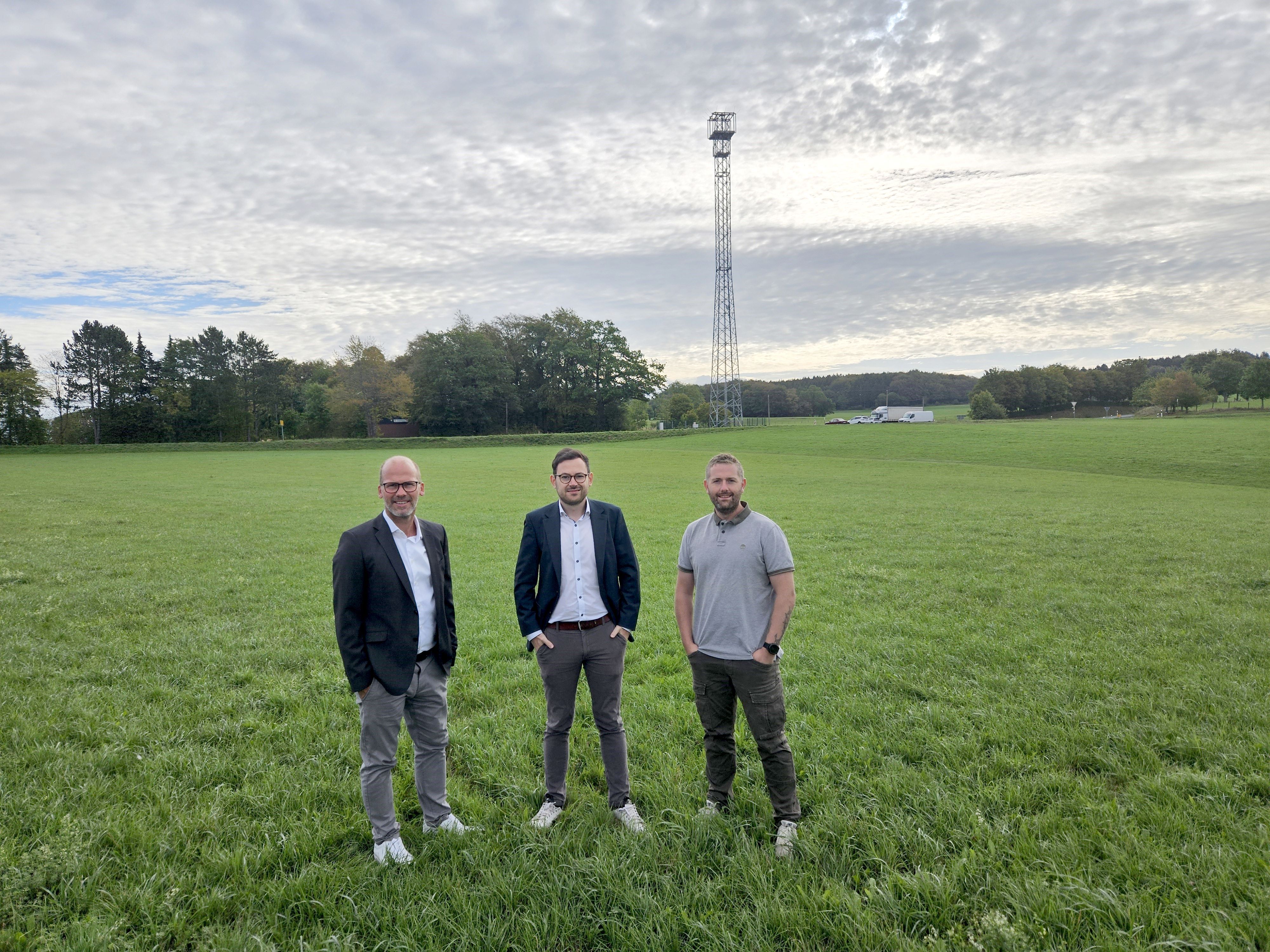 Drei Männer stehen nebeneinander auf einer Wiese. Im Hintergrund ist der Funkmast zu sehen.
