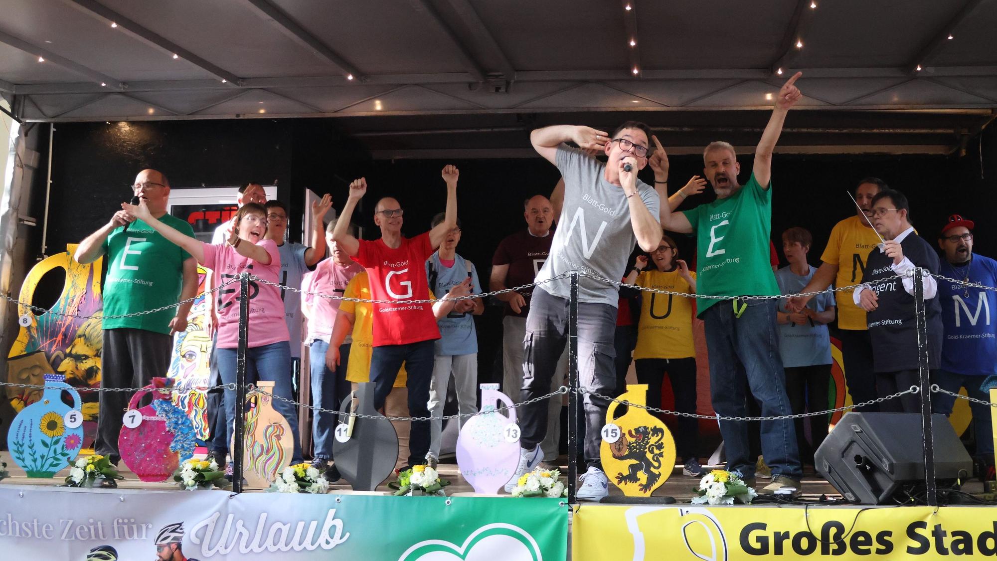Auf dem Bild sind Chormitglieder in bunten T-Shirts auf der Bühne zu sehen.