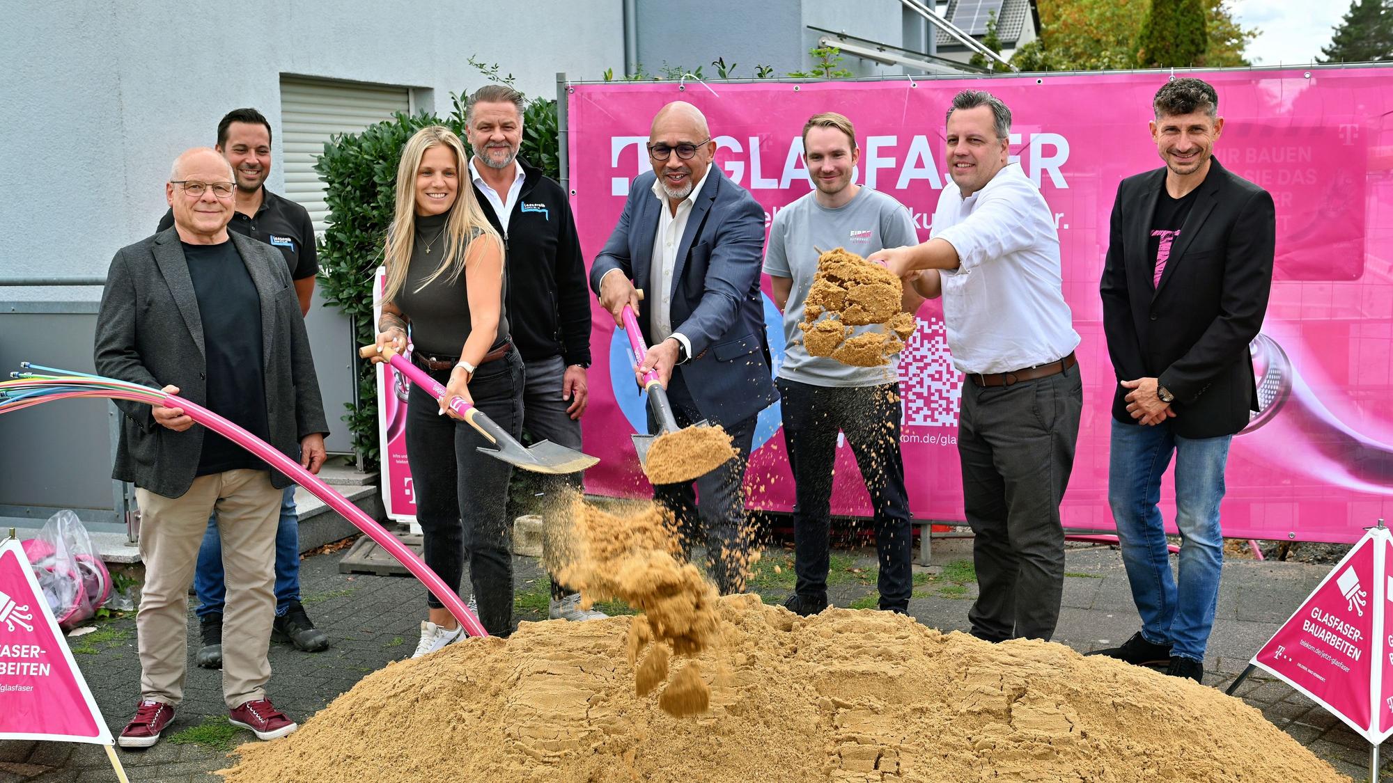 Zu sehen ist eine Frau und sieben Männer vor einem Sandhaufen. Die Frau und zwei Männer halten eine Spaten mit Sand in die Höhe.