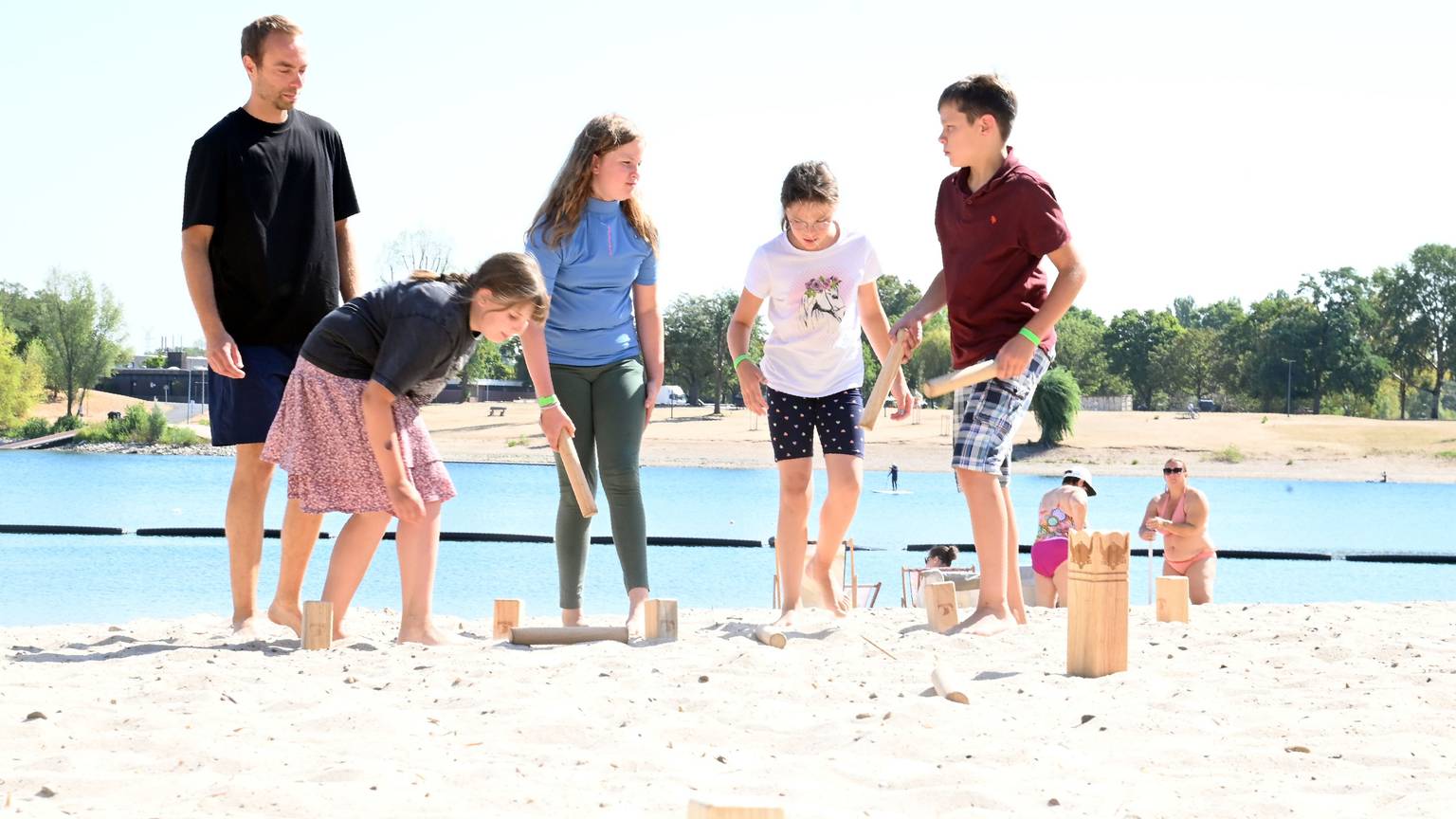 Ein Mitarbeiter des Vereins Bunter Kreis Rheinland e.V. steht mit vier Geschwistern von schwerkranken Kindern am Strand des Fühlinger Sees und spielt mit ihnen Wikingerschach.