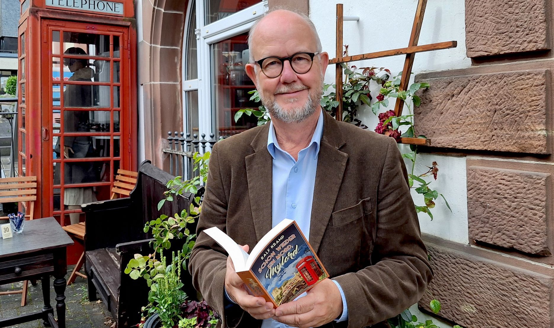 Der Krimi-Autor Ralf Kramp steht vor einer Hauswand und hält sein neues Buch in Händen.