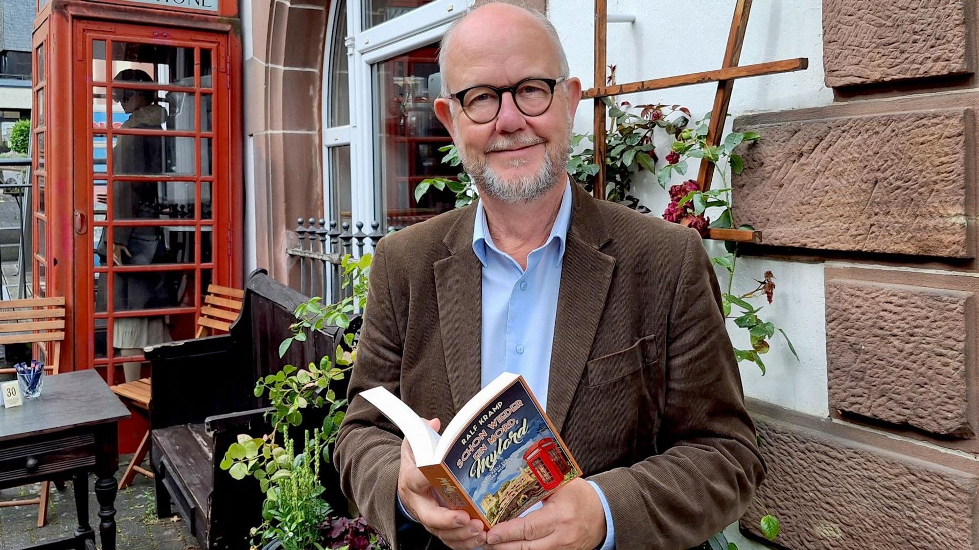 Der Krimi-Autor Ralf Kramp steht vor einer Hauswand und hält sein neues Buch in Händen.