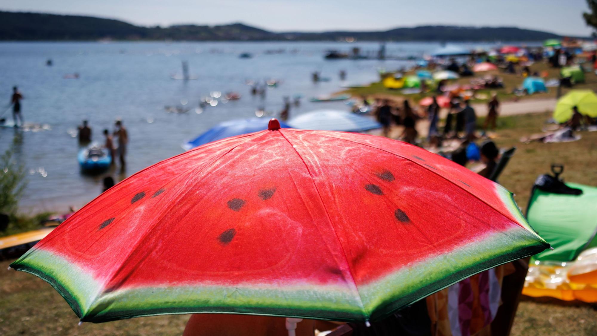 Ein Sonnenschirm mit dem Aufdruck einer Wassermelone steht am Sonntag bei heißem Wetter am gut besuchten Badestrand am Brombachsee.