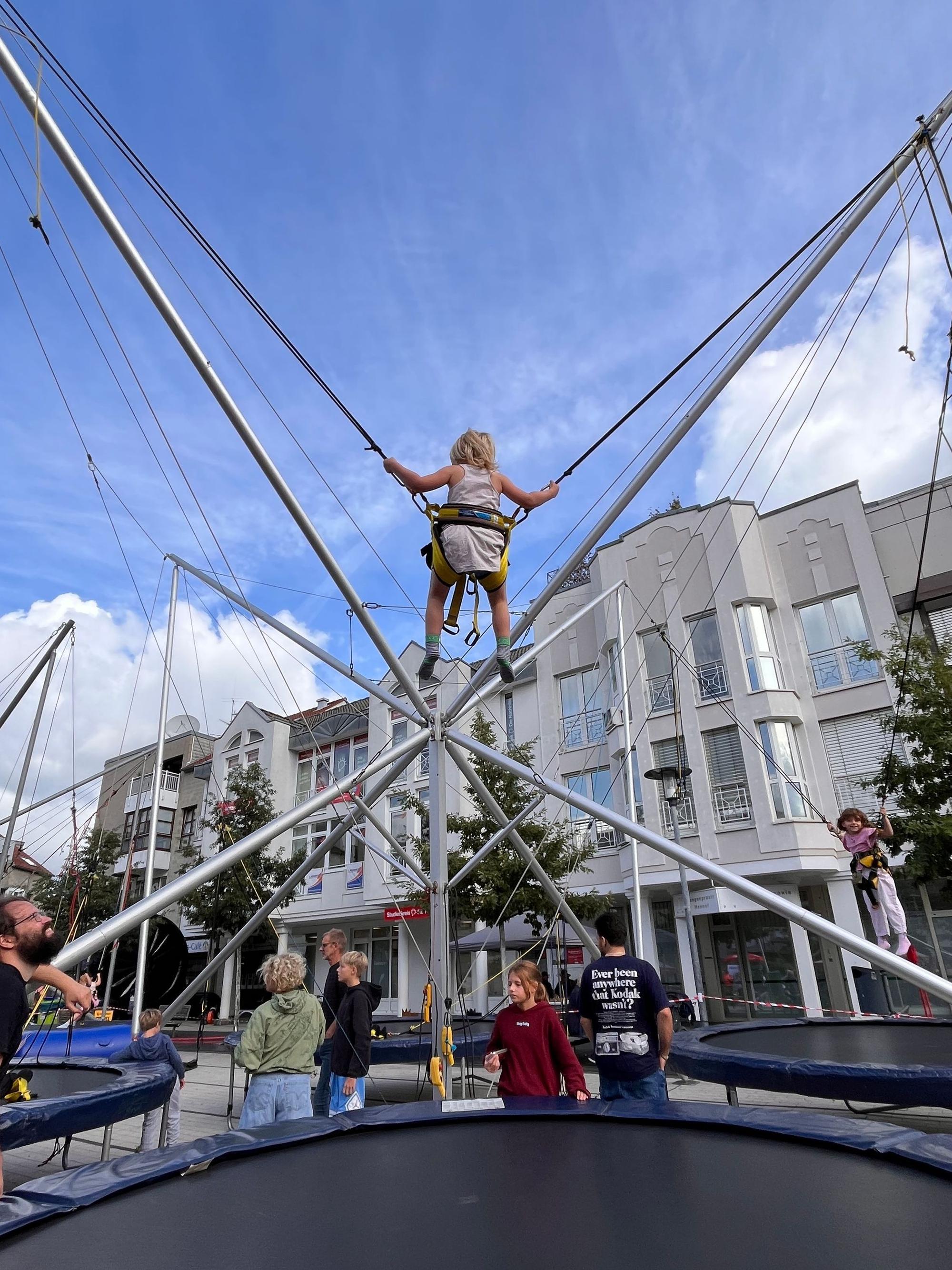 Ein kleines Mädchen schwingt in einem Tragesitz über einem Trampolin auf und ab.