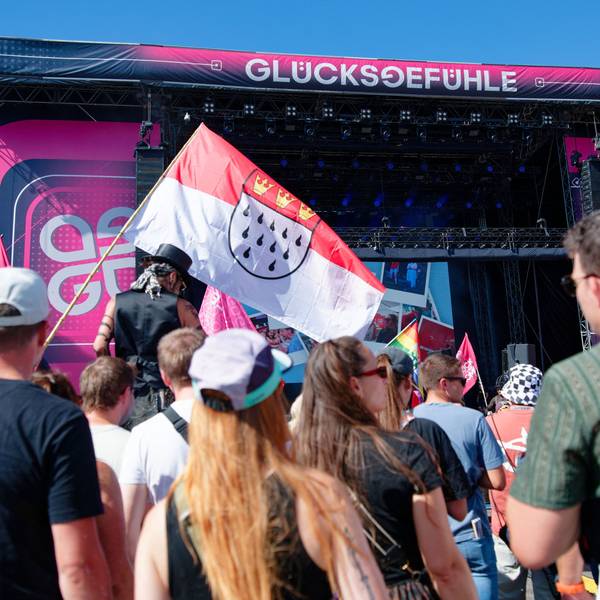 Besucher stehen vor einer Bühne beim Glücksgefühle-Festival auf dem Hockenheimring und halten eine Kölner Stadtflagge. Auch der Kölner Männerchor „Die Gürngürtelrosen“ trat auf dem Hockenheimring auf.