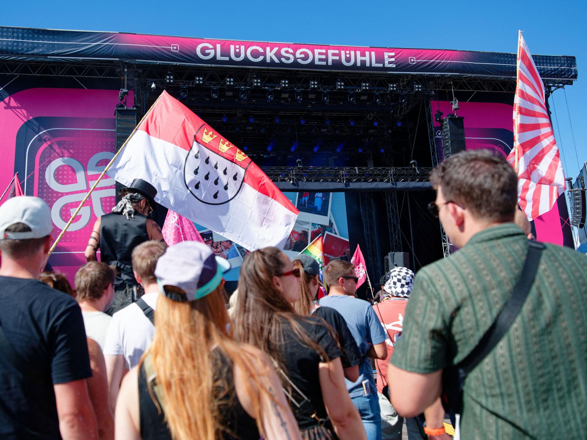Besucher stehen vor einer Bühne beim Glücksgefühle-Festival auf dem Hockenheimring und halten eine Kölner Stadtflagge. Auch der Kölner Männerchor „Die Gürngürtelrosen“ trat auf dem Hockenheimring auf.