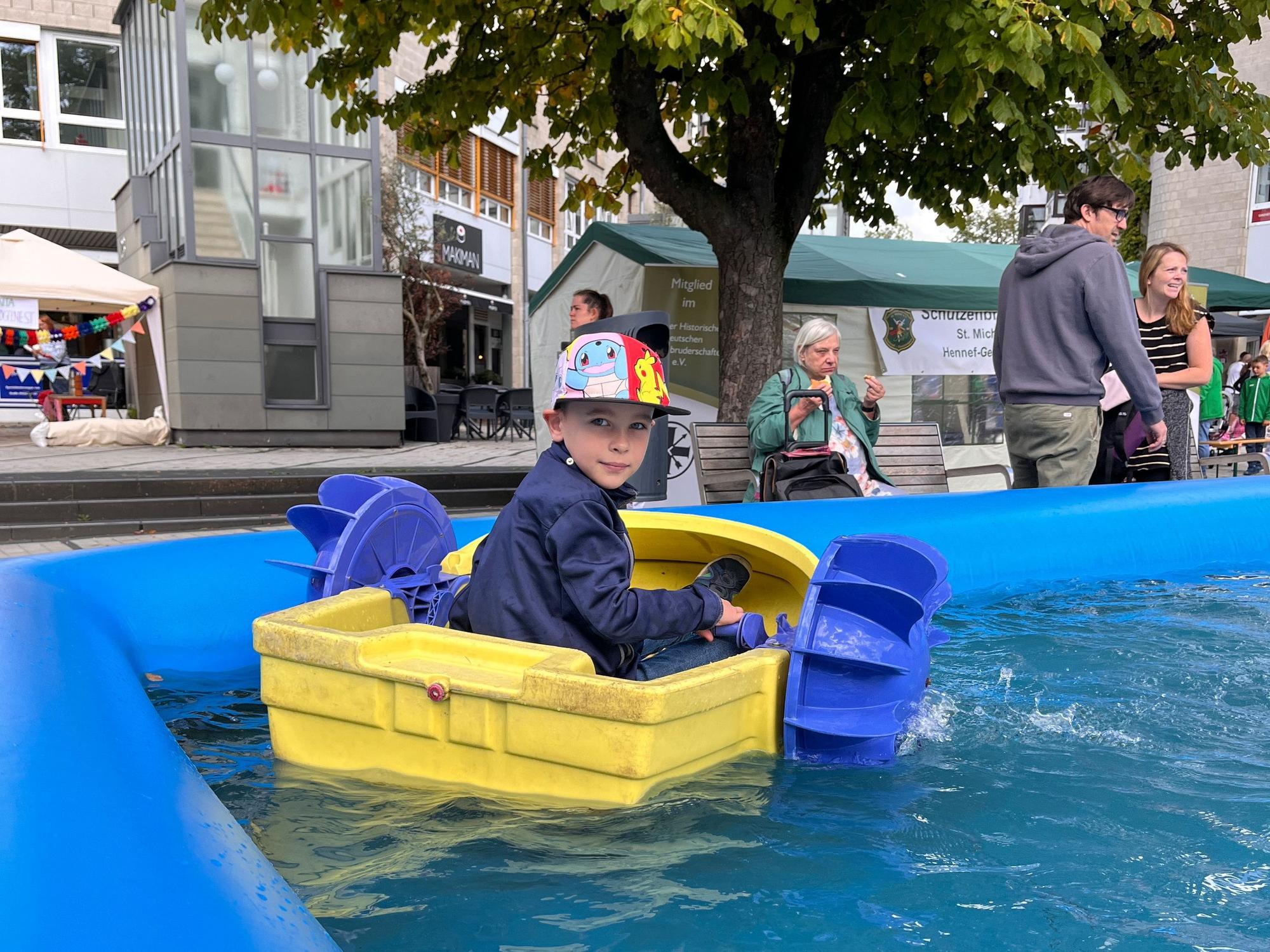 Ein Junge mit Baseballkappe sitzt in einem kleinen Boot mit Schaufelrädern, das in einem Wasserbecken schwimmt.