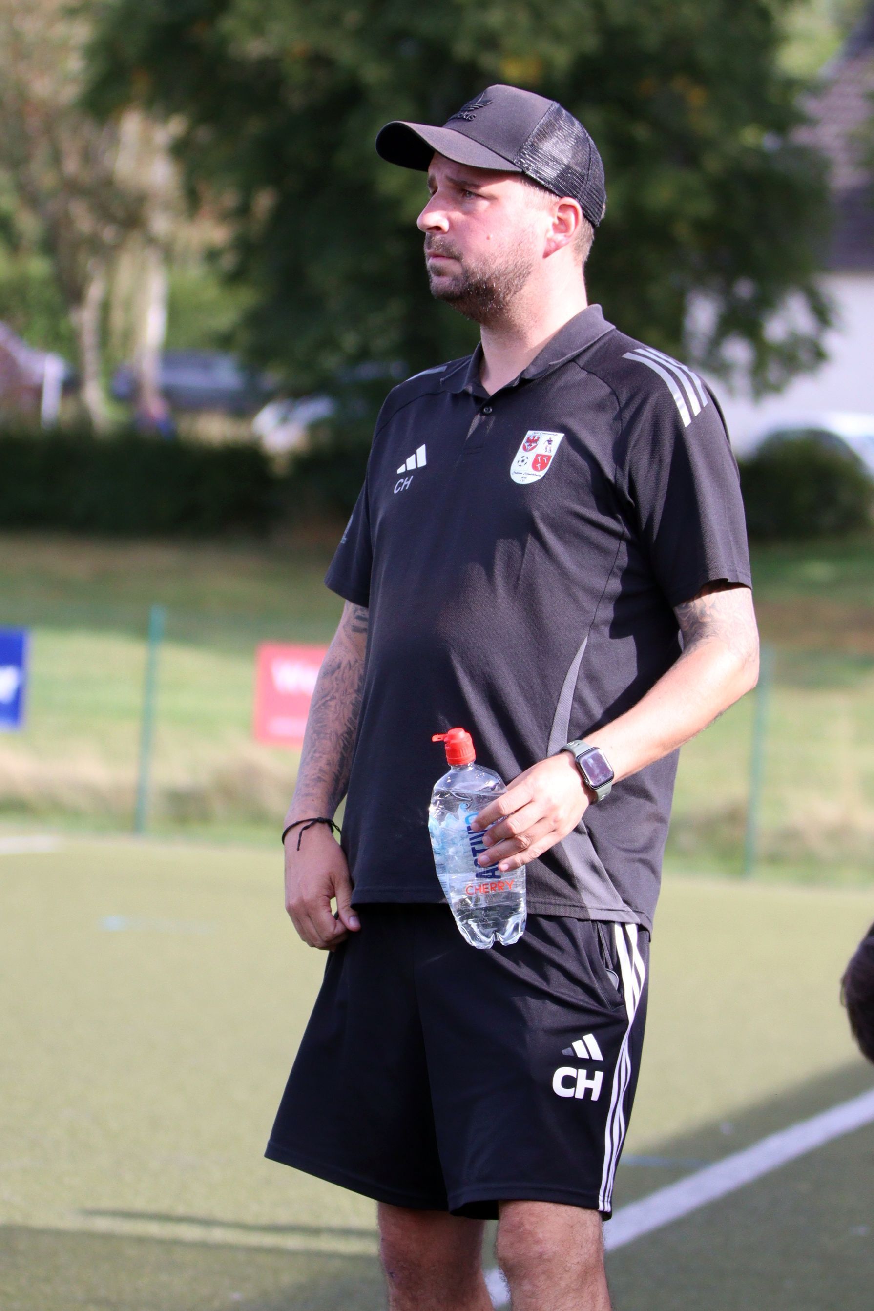 Dahlems Trainer Christian Hammes hält in einer Hand eine Wasserflasche und schaut dem Spiel zu.
