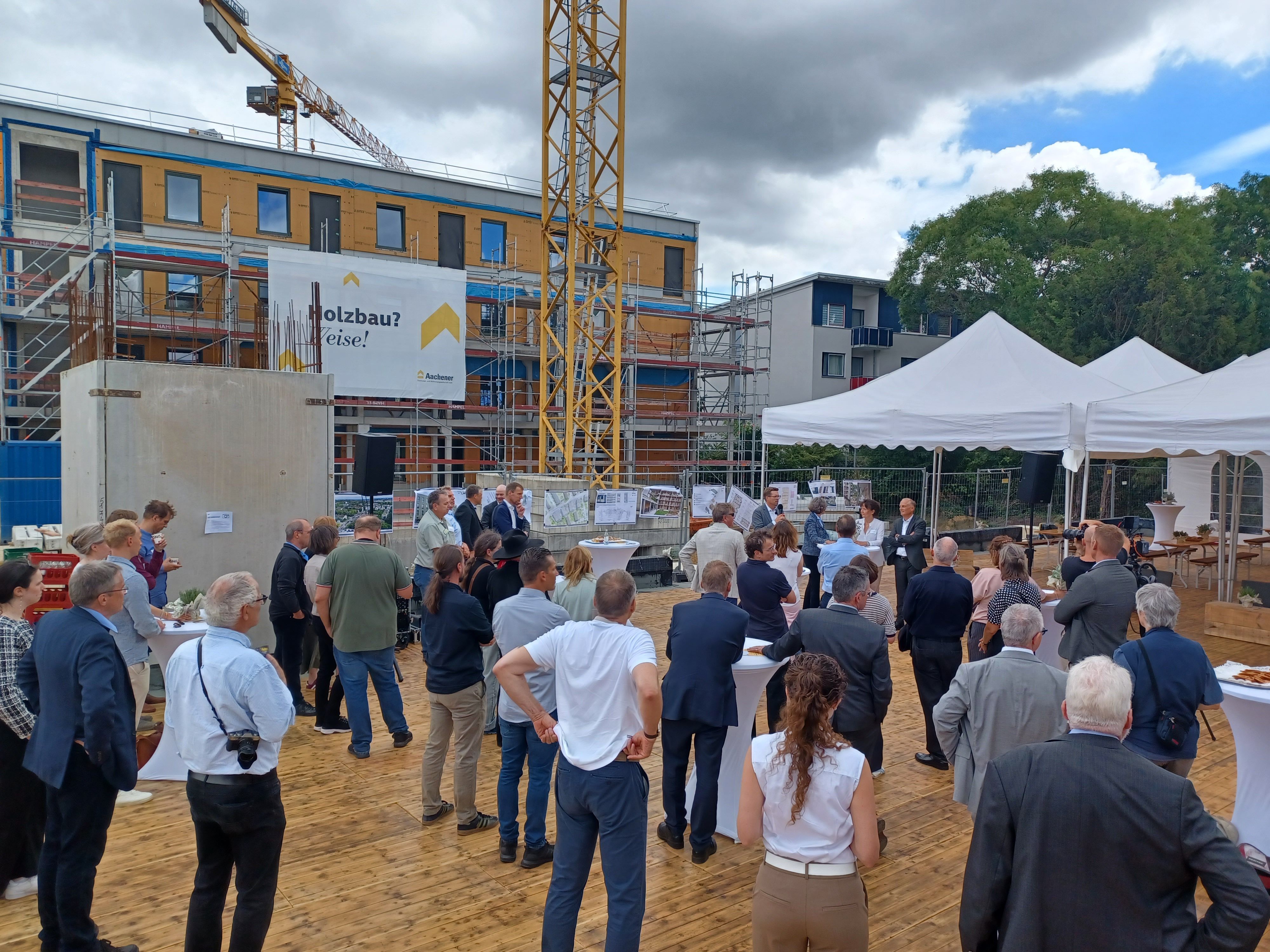 Viele Menschen stehen auf einer Baustelle. Im Hintergrund sind ein eingerüsteter Rohbau und ein Kran zu sehen.