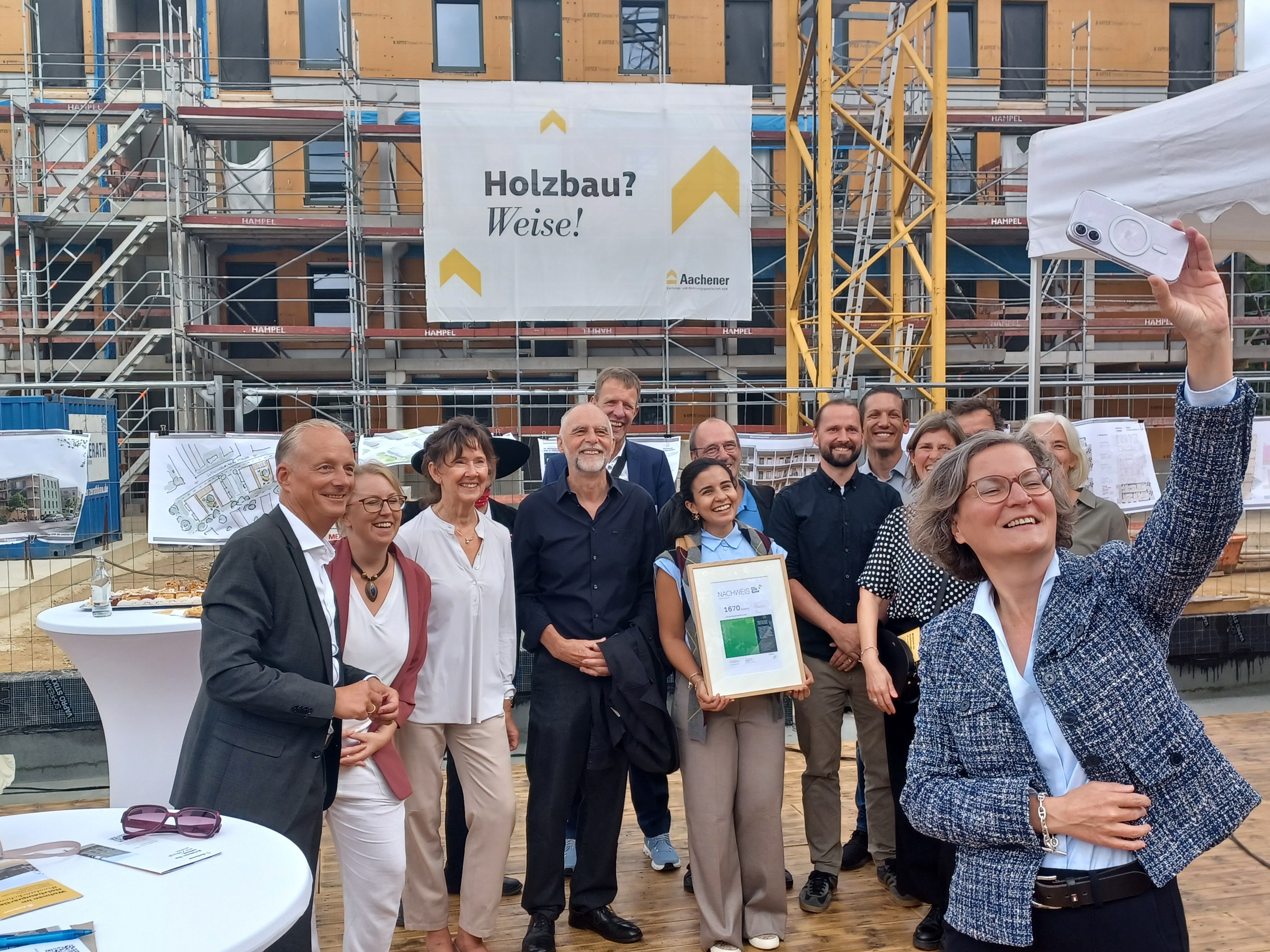 Eine Menschengruppe steht vor einem Rohbau. Eine Frau mit kinnlangem Haar macht ein Selfie.