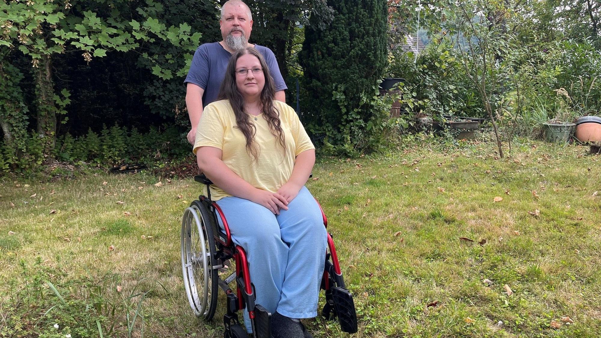 Das junge Mädchen sitzt im Rollstuhl, der Vater steht dahinter.