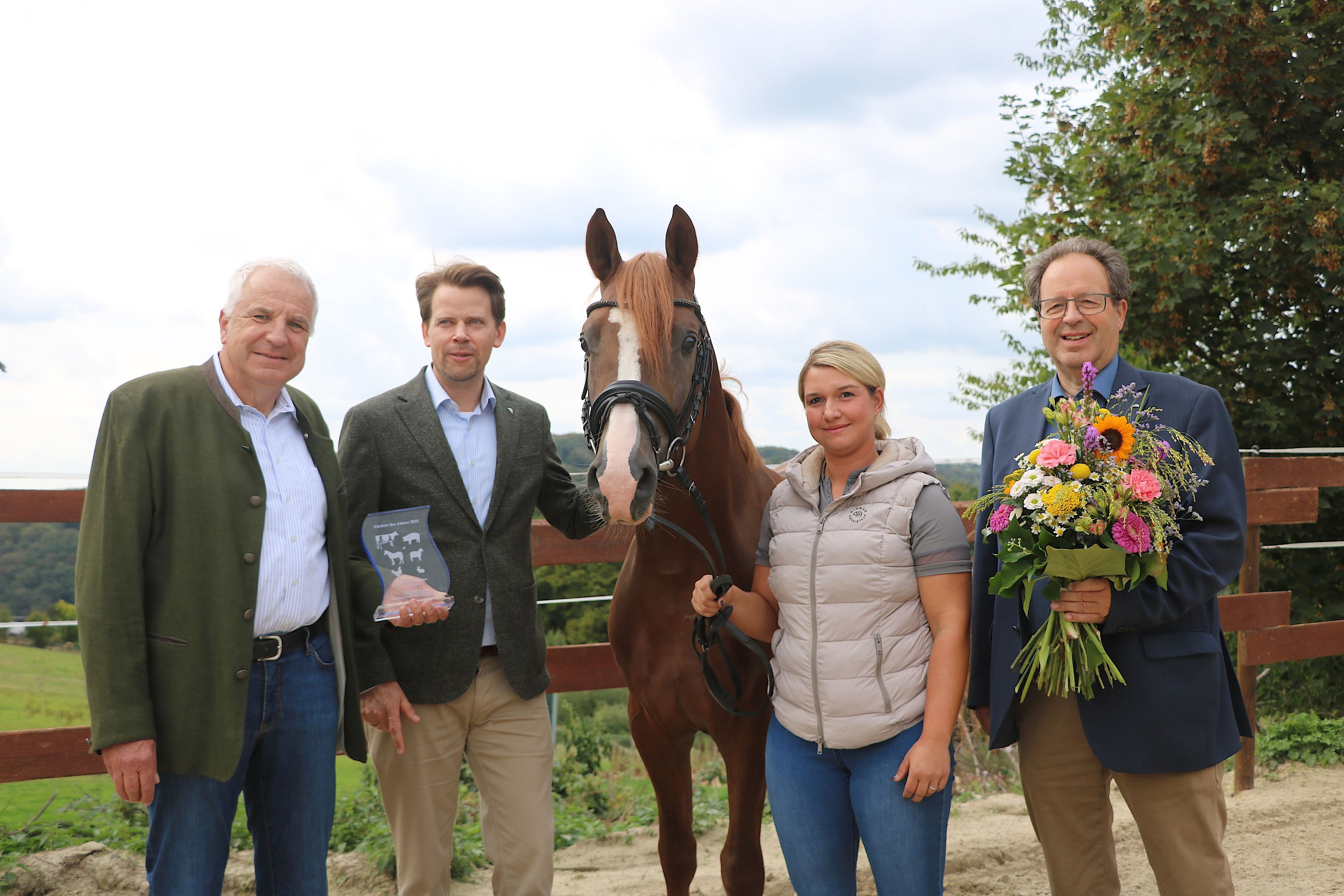 Eine blonde Frau hält ein braunes Pferd am Zügel, daneben stehen drei Männer, einer mit Blumenstrauß.