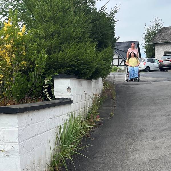 Die junge Frau wird im Rollstuhl von ihrer Mutter geschoben.