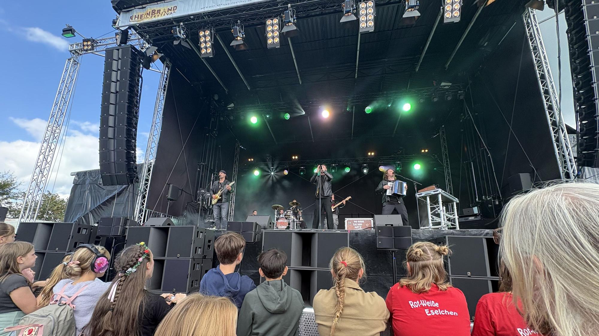 Jugendliche stehen vor einer großen Bühne. Dort spielt eine Band.