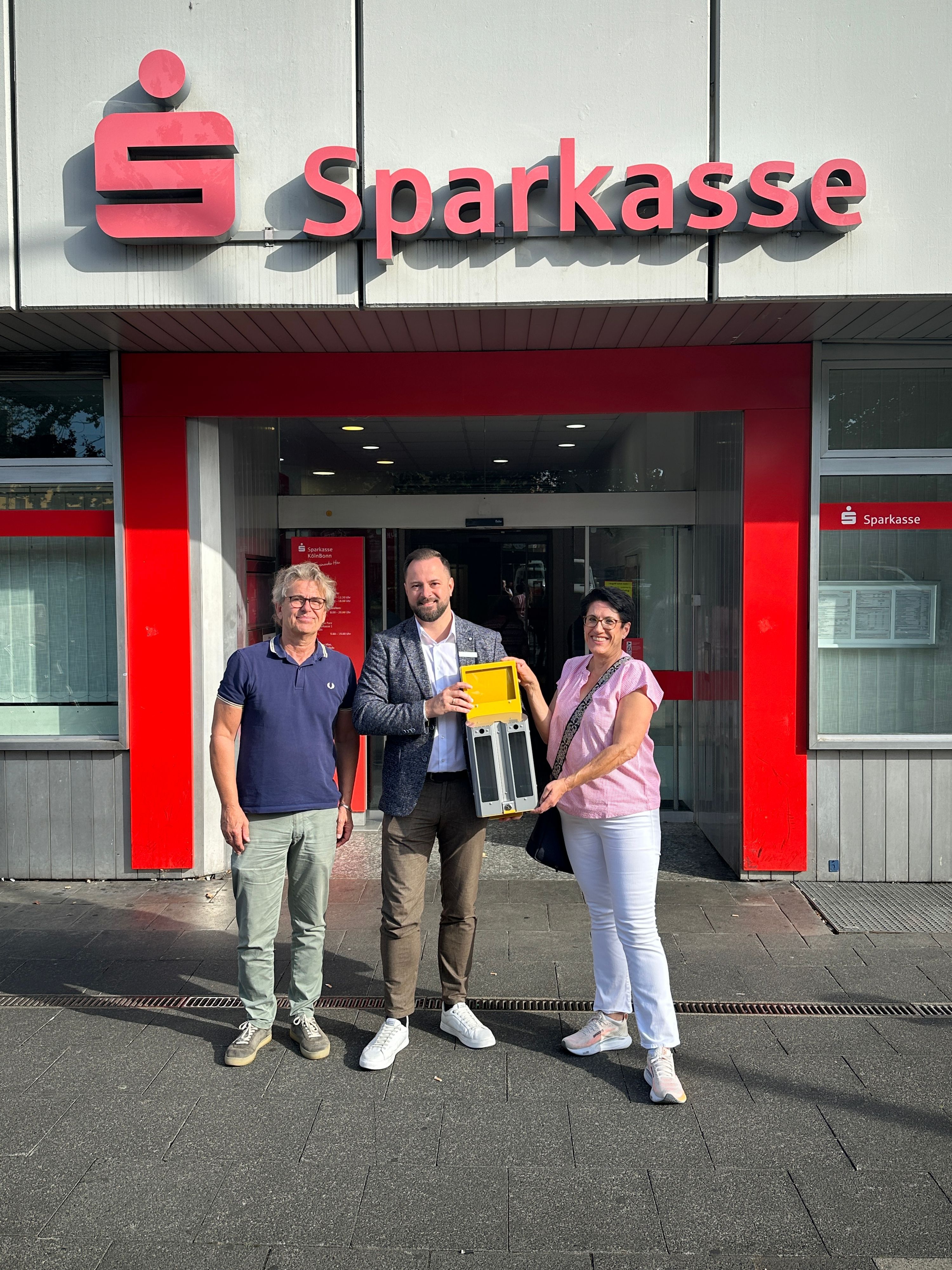 Zwei Männer und eine Frau stehen vor einer Sparkassen-Filiale.