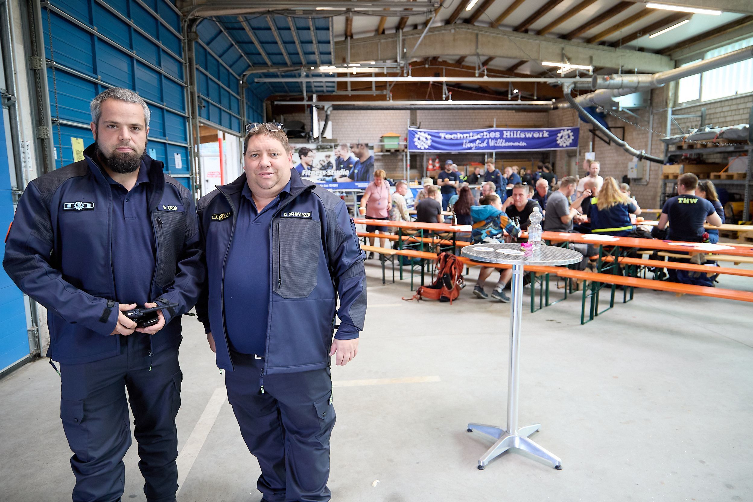Björn Gross und Daniel Schwarzer stehen in THW-Jacken nebeneinander in einer Halle.