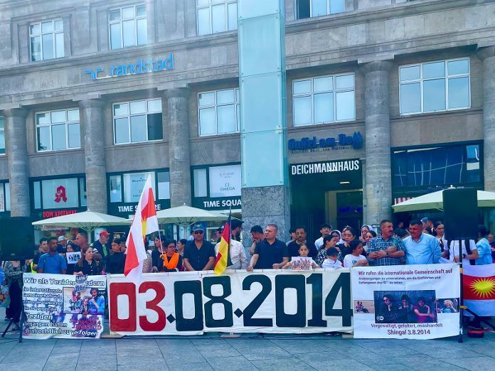 Menschen stehen hinter einem Banner, das an das Massaker an den Jesiden am 3.8. 2014 erinnert.