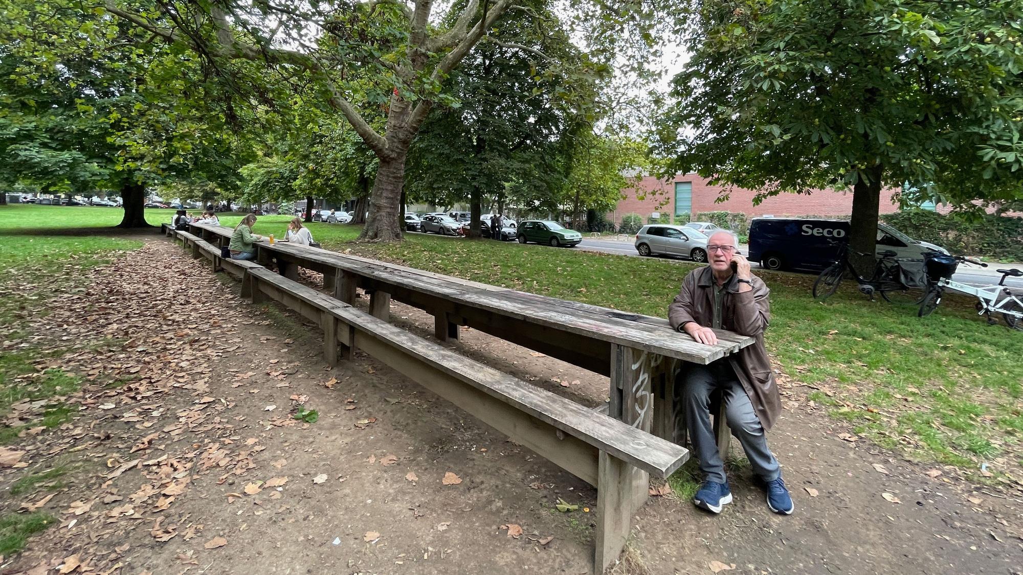 Ein Mann sitzt an einem sehr langen Holztisch in einer Grünanlage.